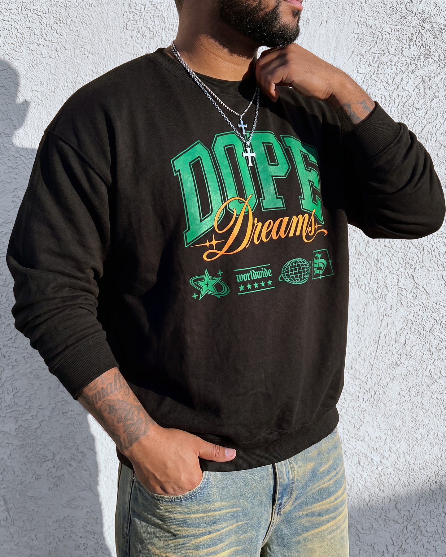 A man wearing a black crewneck sweatshirt with a green and gold graphic print that reads 'DOPE Dreams' and light wash blue jeans, standing against a white textured wall.