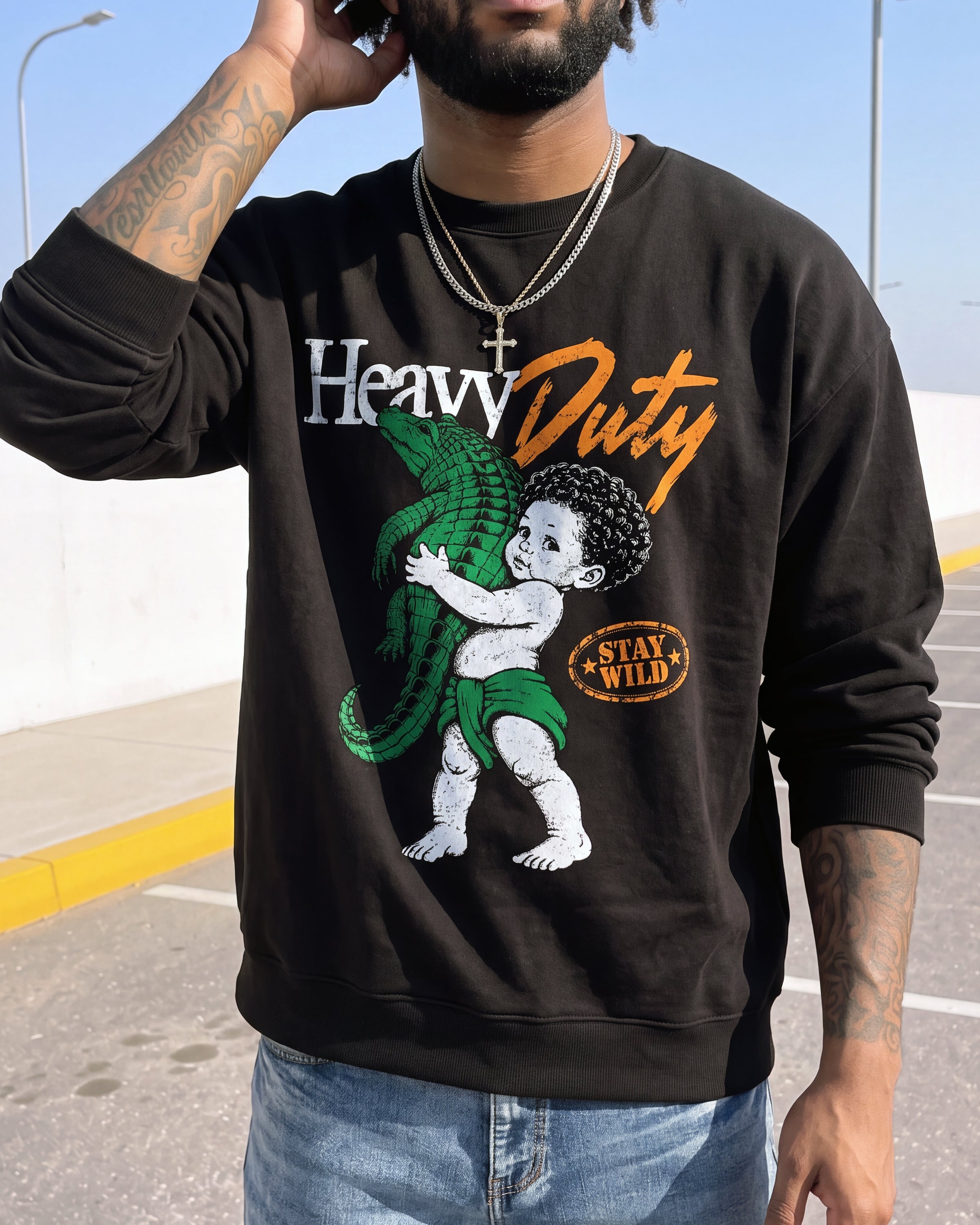A man wearing a black crewneck sweatshirt with a graphic print of a baby holding a green alligator, with the text 'Heavy Duty' and 'STAY WILD'.