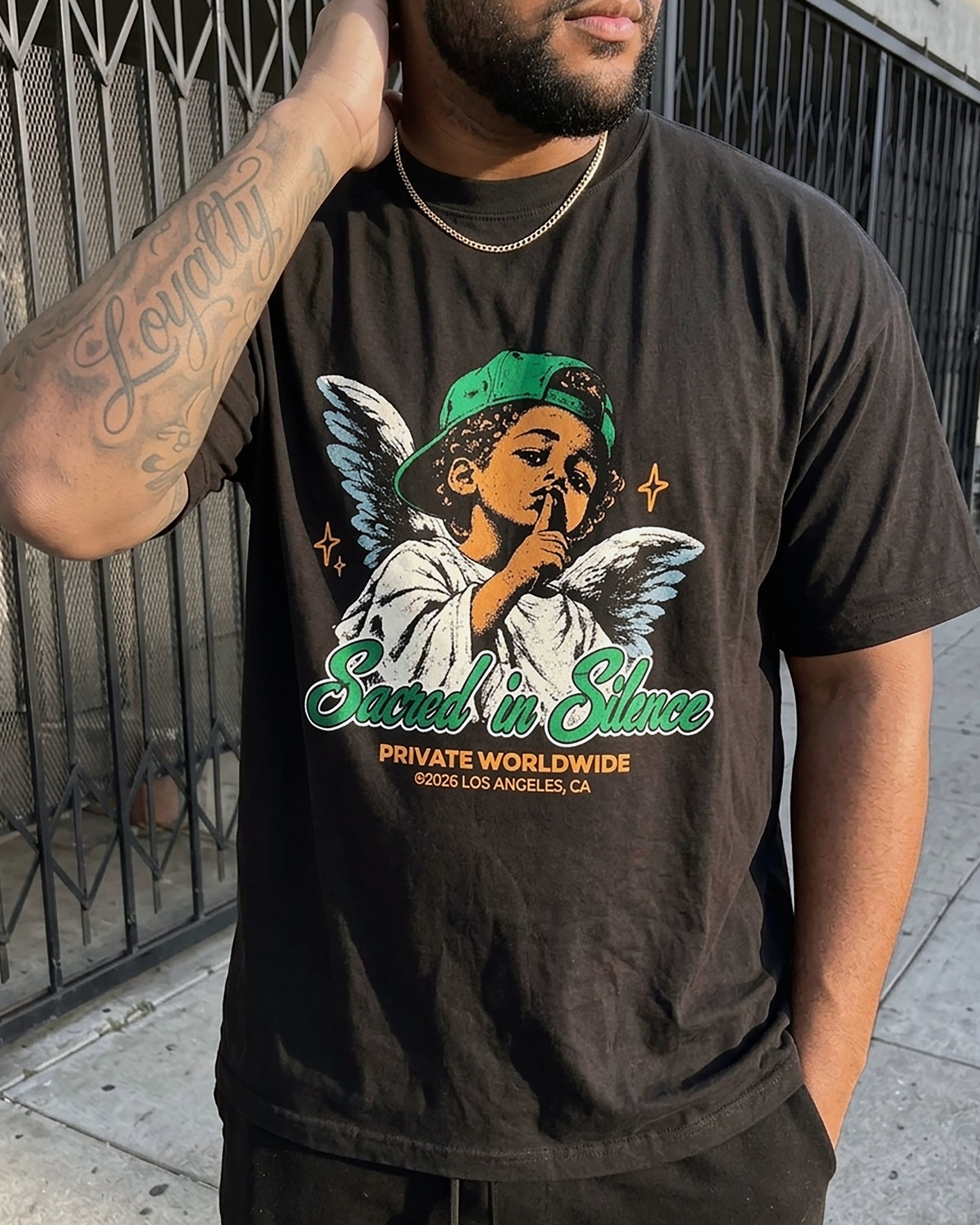 A man wearing a black graphic t-shirt with a colorful illustration of an angel and the text 'Sacred in Silence' and 'PRIVATE WORLDWIDE LOS ANGELES, CA'.