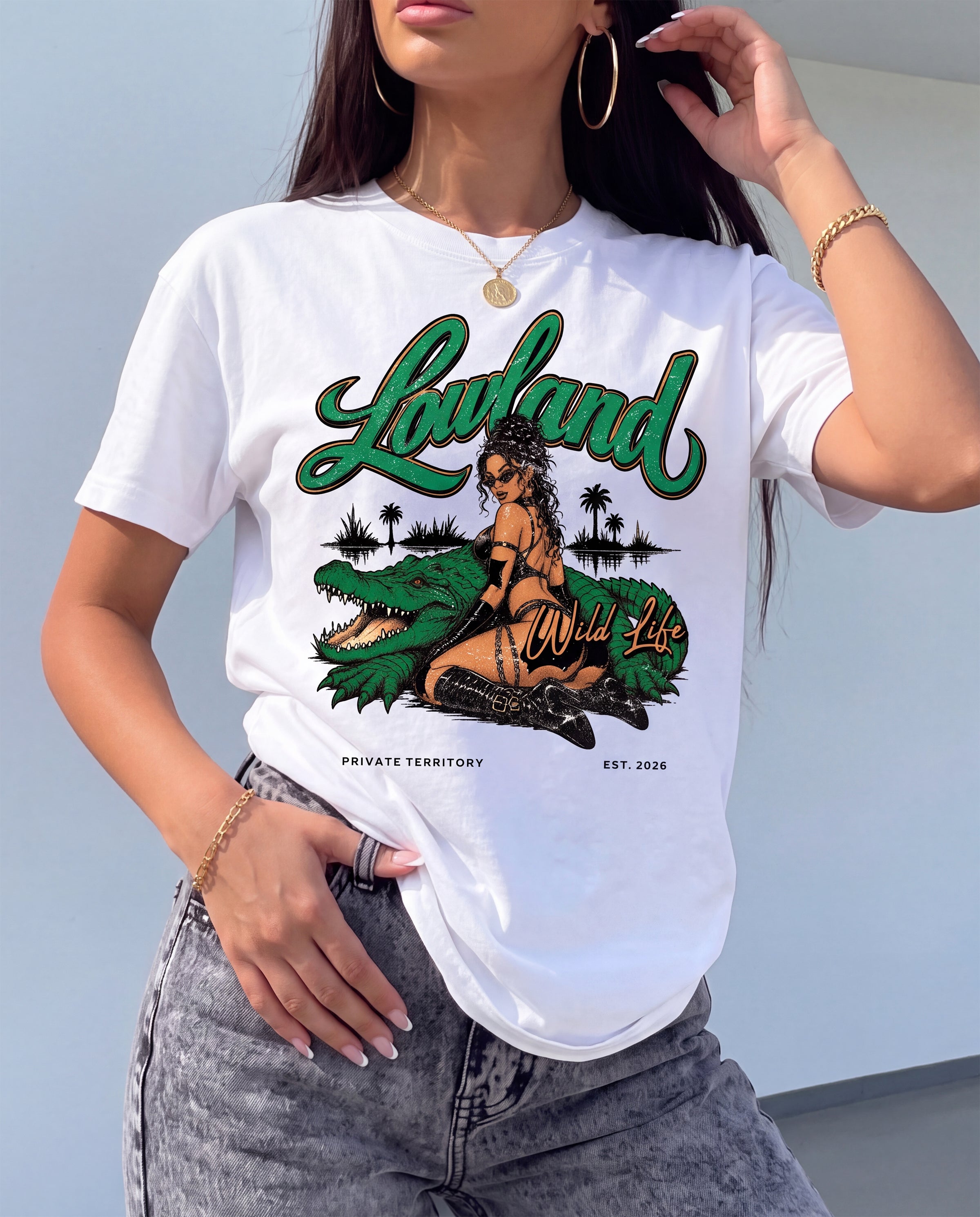 A woman wearing a white graphic t-shirt with a green and black 'Lowland Wild Life' print featuring a crocodile and a woman, paired with gray denim jeans.