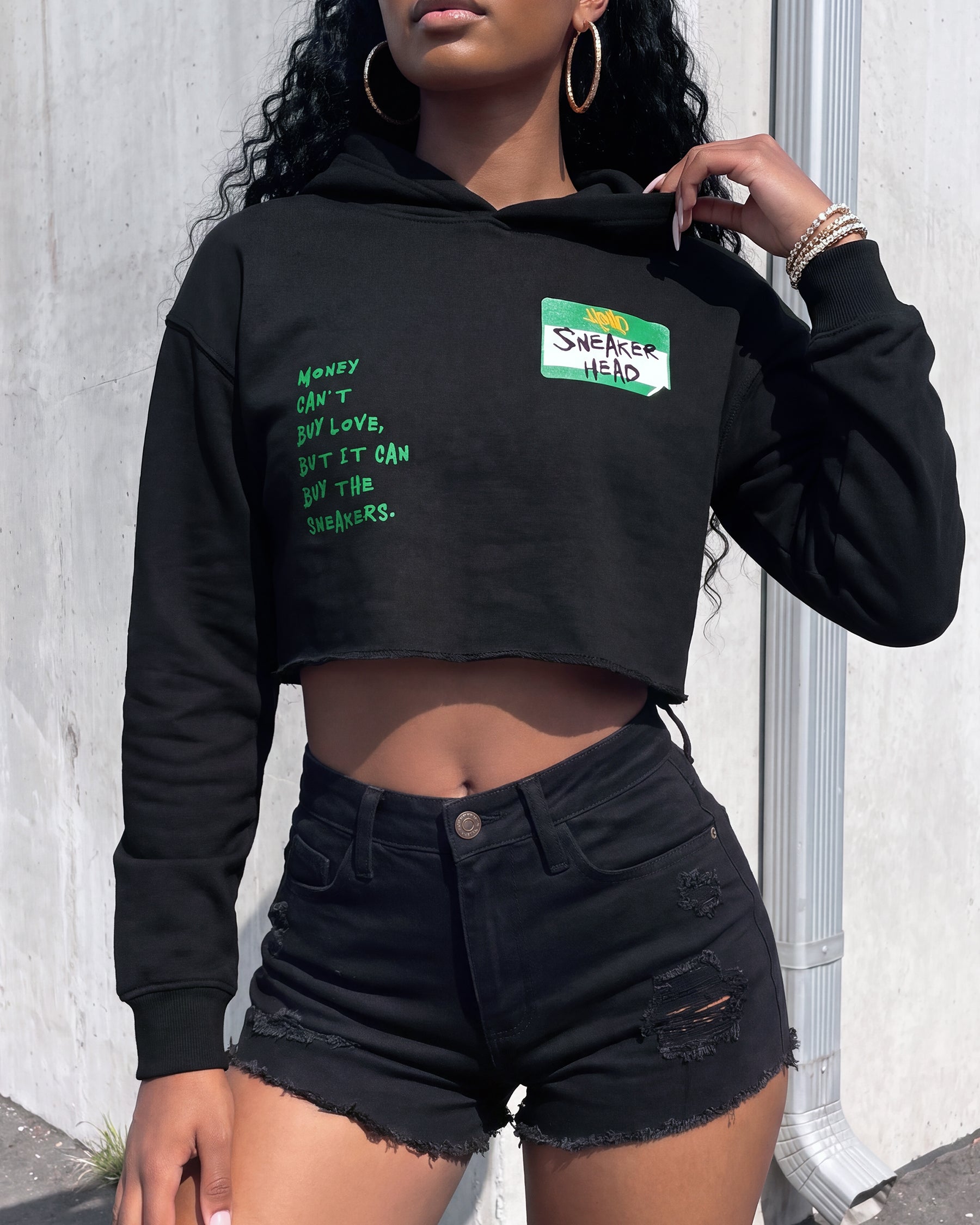 A woman wearing a black cropped hoodie with green text on the left chest and a small green-and-white 'SHE'S HERE' patch on the right chest, paired with black distressed shorts, standing against a light gray wall.