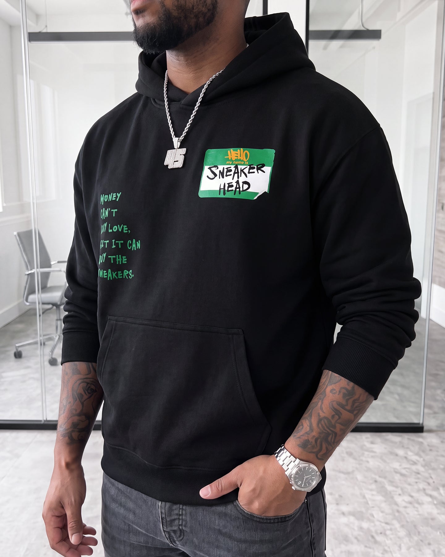 A man wearing a black hoodie with a green and white 'Hello SNEAKER HEAD' name-tag graphic on the chest, paired with gray jeans in a modern office setting.
