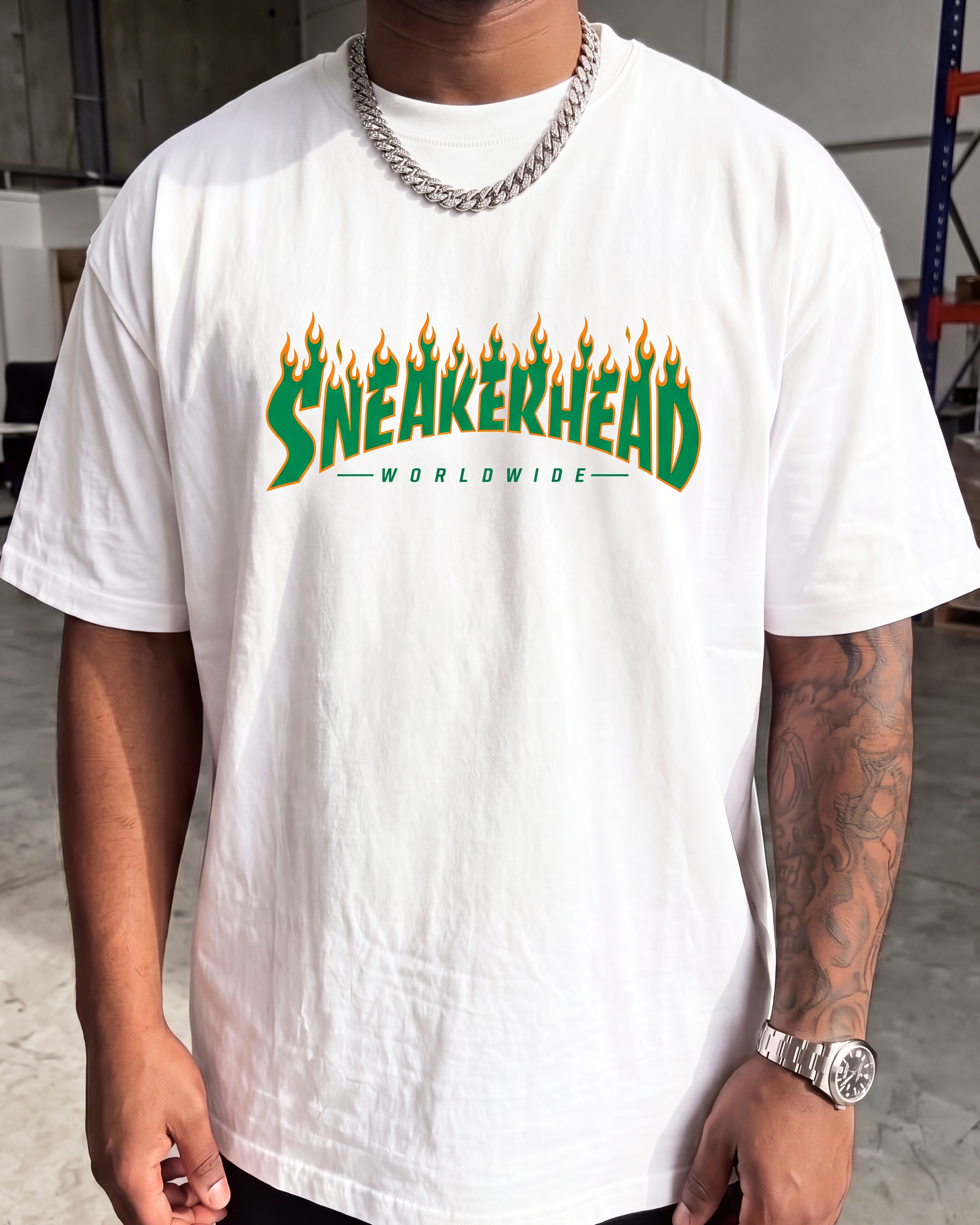 A man wearing a white crewneck t-shirt with a green and yellow flame-style 'SNEAKERHEAD' graphic on the chest, paired with a silver chain necklace and a silver watch.