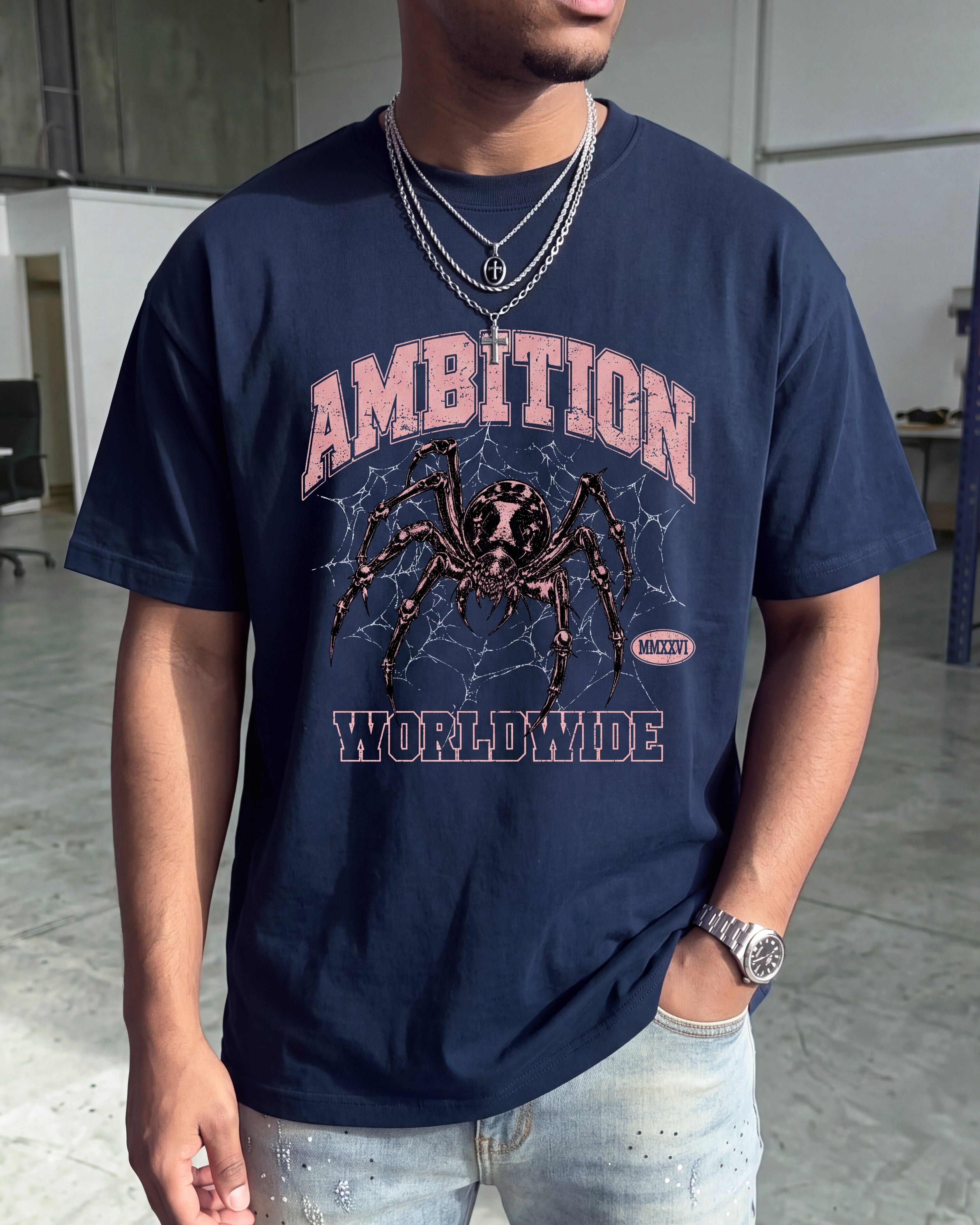 A man wearing a navy blue graphic t-shirt with a spider web and the words 'AMBITION WORLDWIDE' in pink and white, paired with light wash jeans.