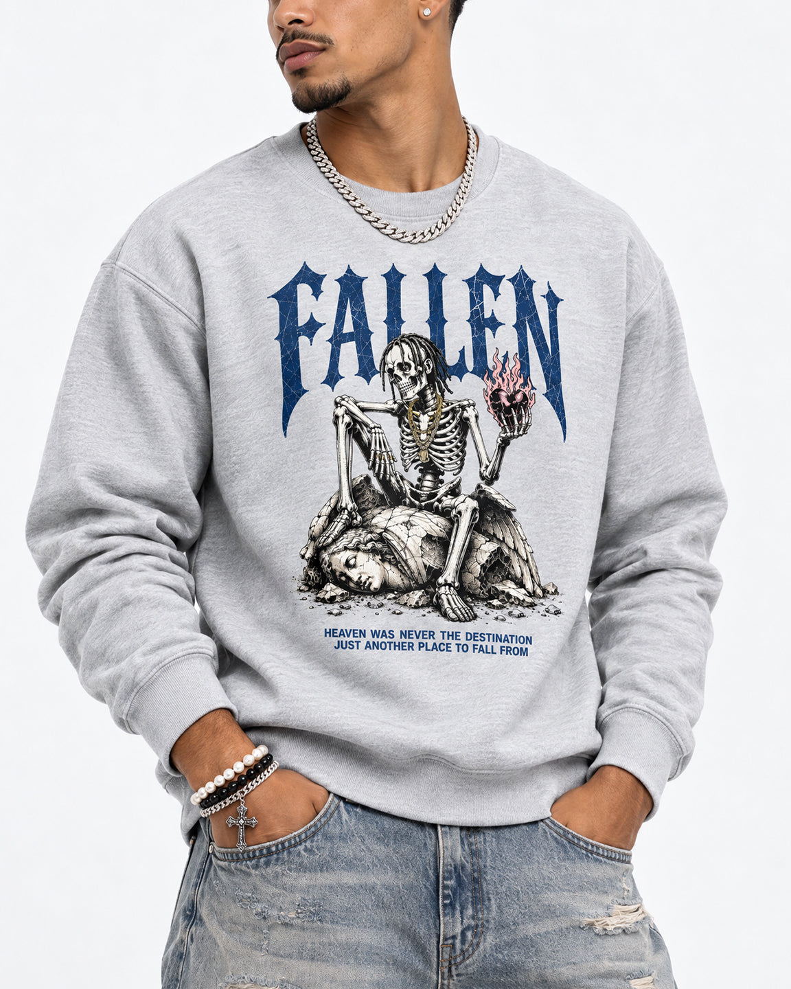 A man wearing a light gray crewneck sweatshirt with a blue and black graphic of a skeleton and the word 'FALLEN' on the front, paired with distressed blue jeans and a silver chain necklace.