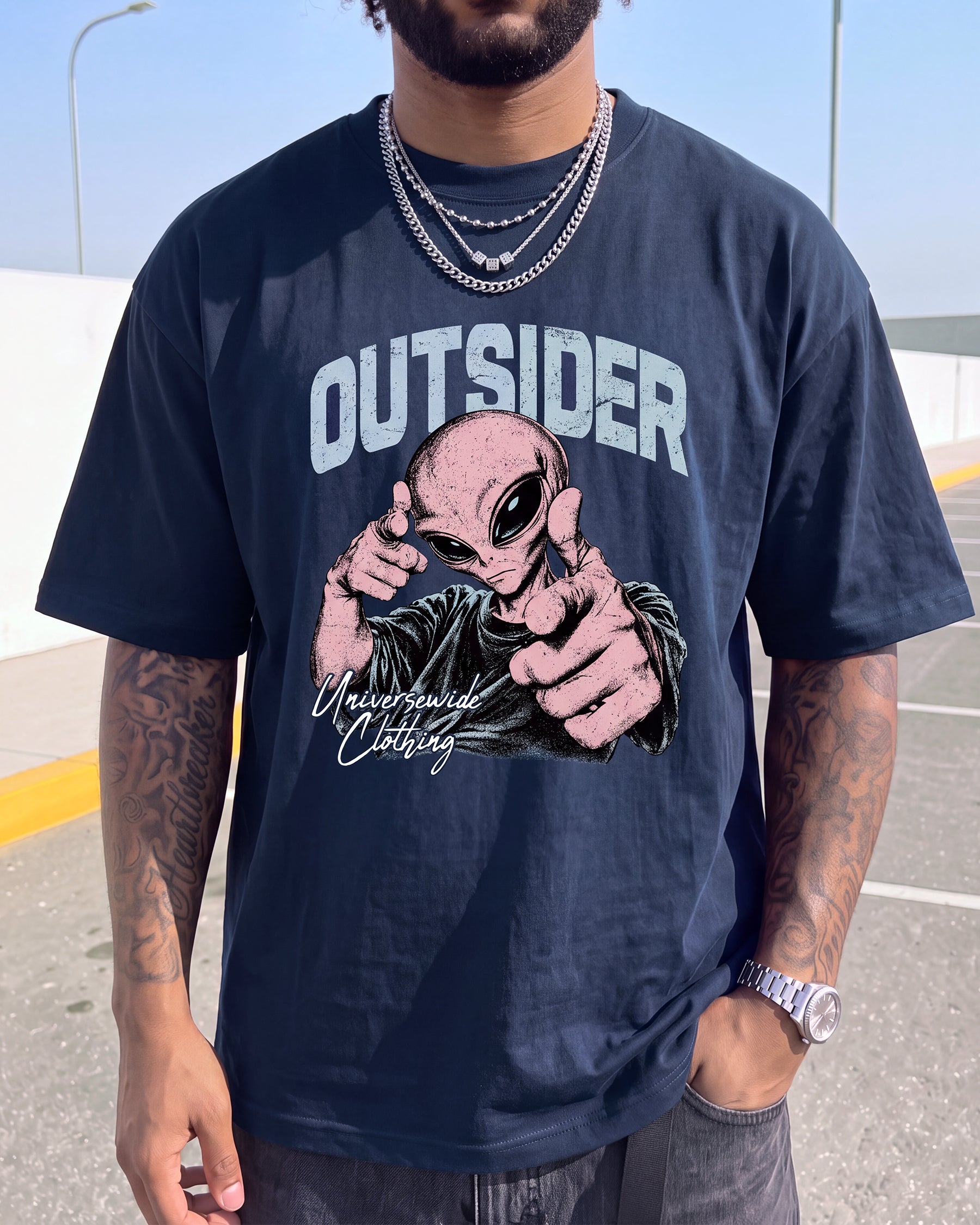 A man wearing a navy blue graphic t-shirt with a pink and black alien design and the word 'OUTSIDER' printed on the front, set against an outdoor background.