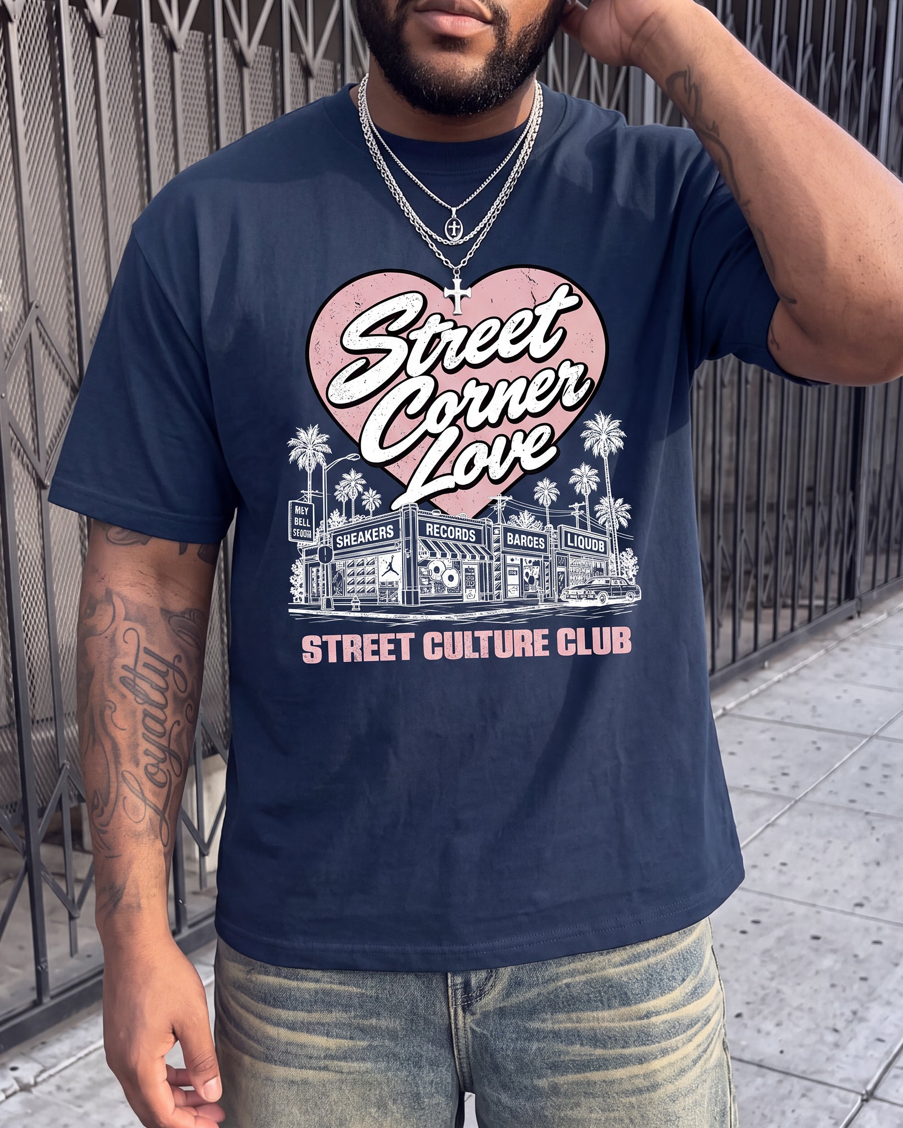 A man wearing a navy blue graphic t-shirt with a pink and white 'Street Corner Love' graphic, paired with faded blue jeans, standing outdoors in front of a metal gate.