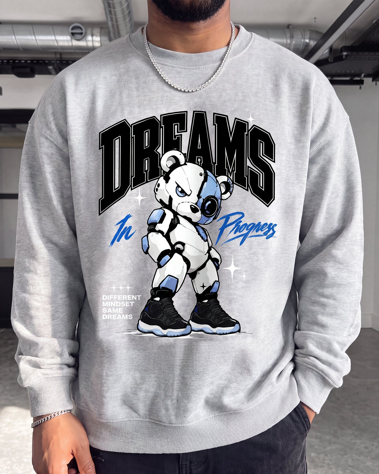 A man wearing a light gray crewneck sweatshirt with a graphic of a white and blue robot bear under the word 'DREAMS' in black arched letters.