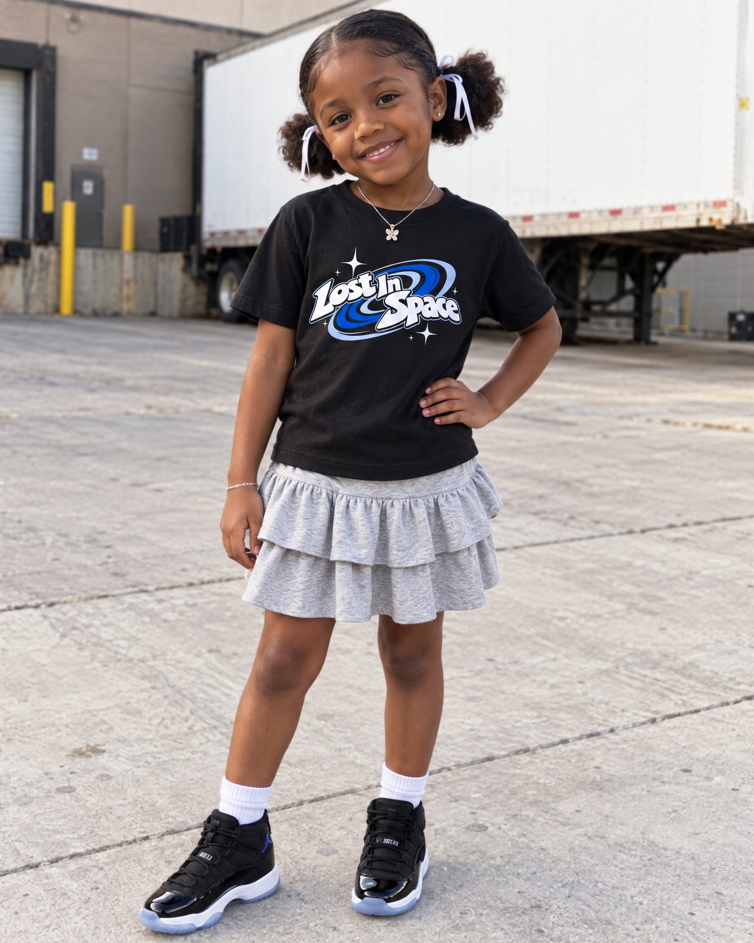 A young girl stands outdoors on a concrete lot, wearing a black graphic T-shirt that reads 'Lost in Space' with blue and white space-themed artwork, a light gray ruffled tiered skirt, white socks, and black sneakers.