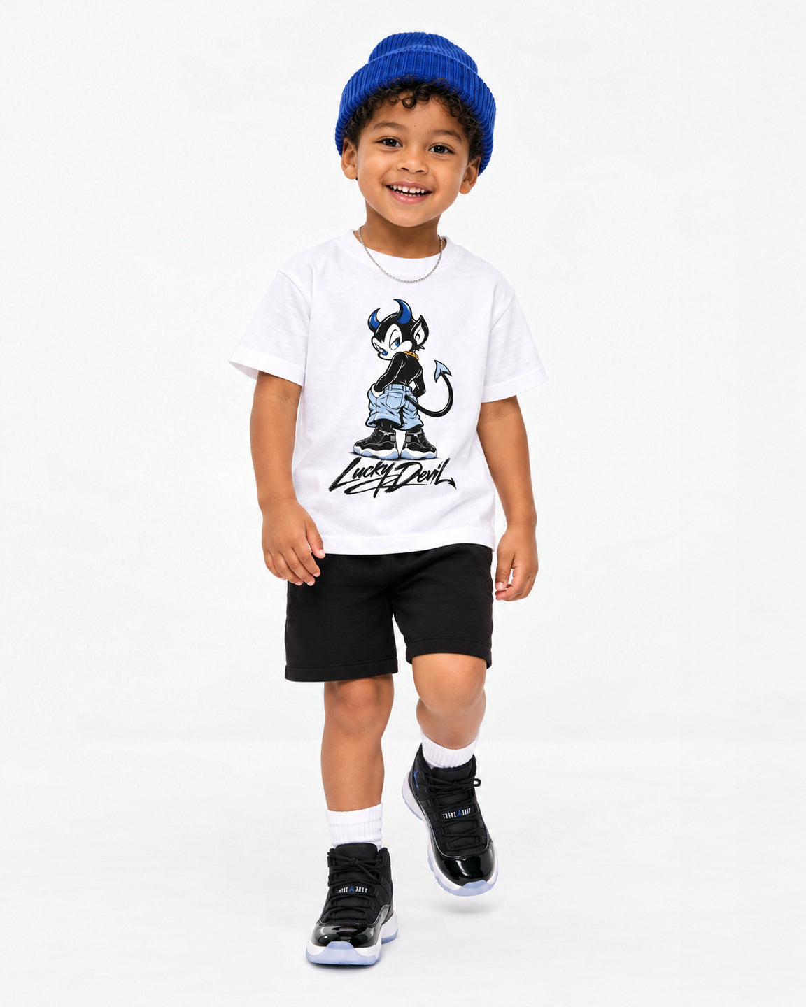 A young boy walking forward in a studio setting, wearing a white graphic t-shirt with a black and blue cartoon character, black shorts, white socks, black sneakers, and a blue knit beanie.
