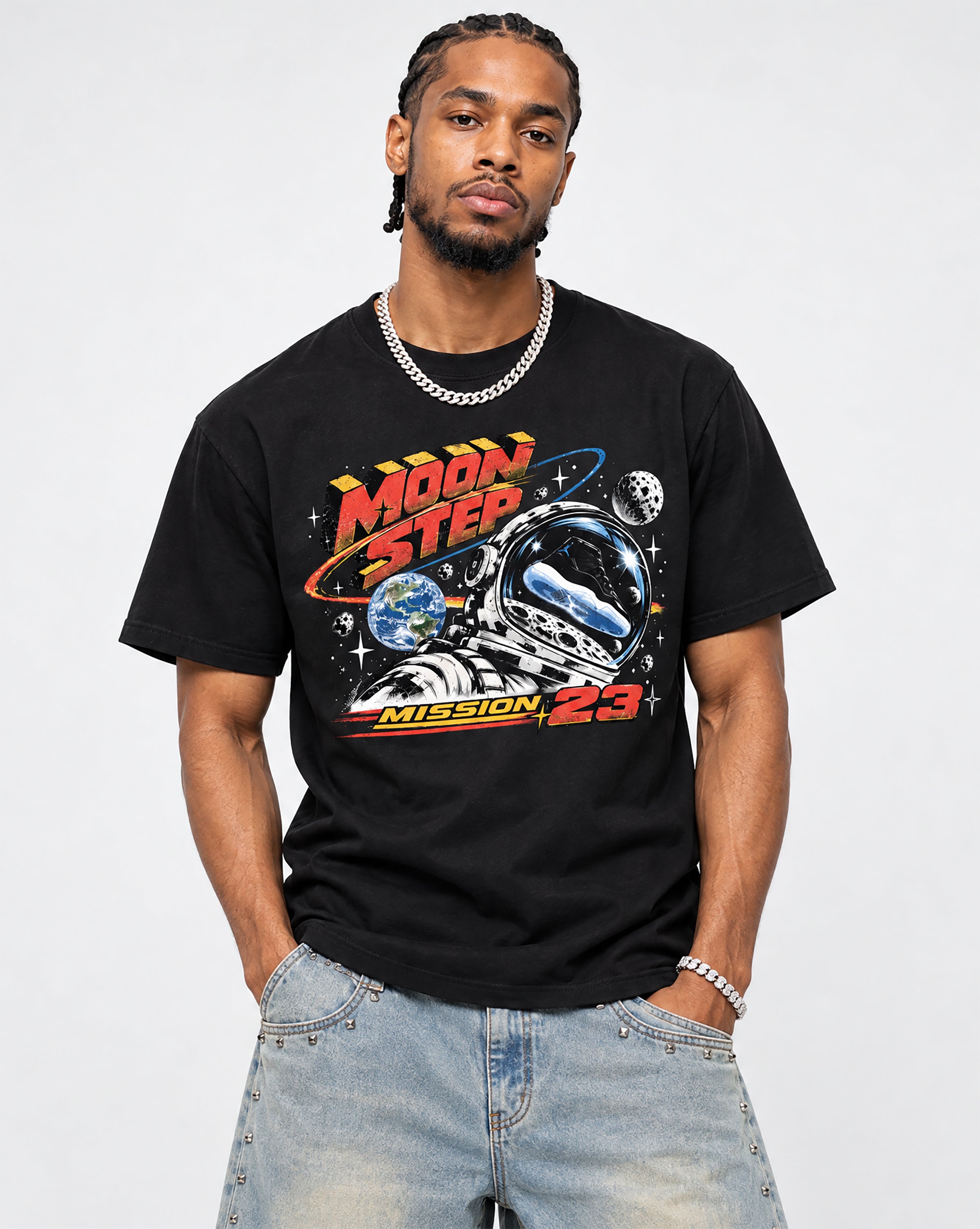 A man wearing a black graphic t-shirt with a space-themed 'Moon Step Mission 23' print, paired with light-wash blue jeans and a silver chain necklace, against a plain light gray background.