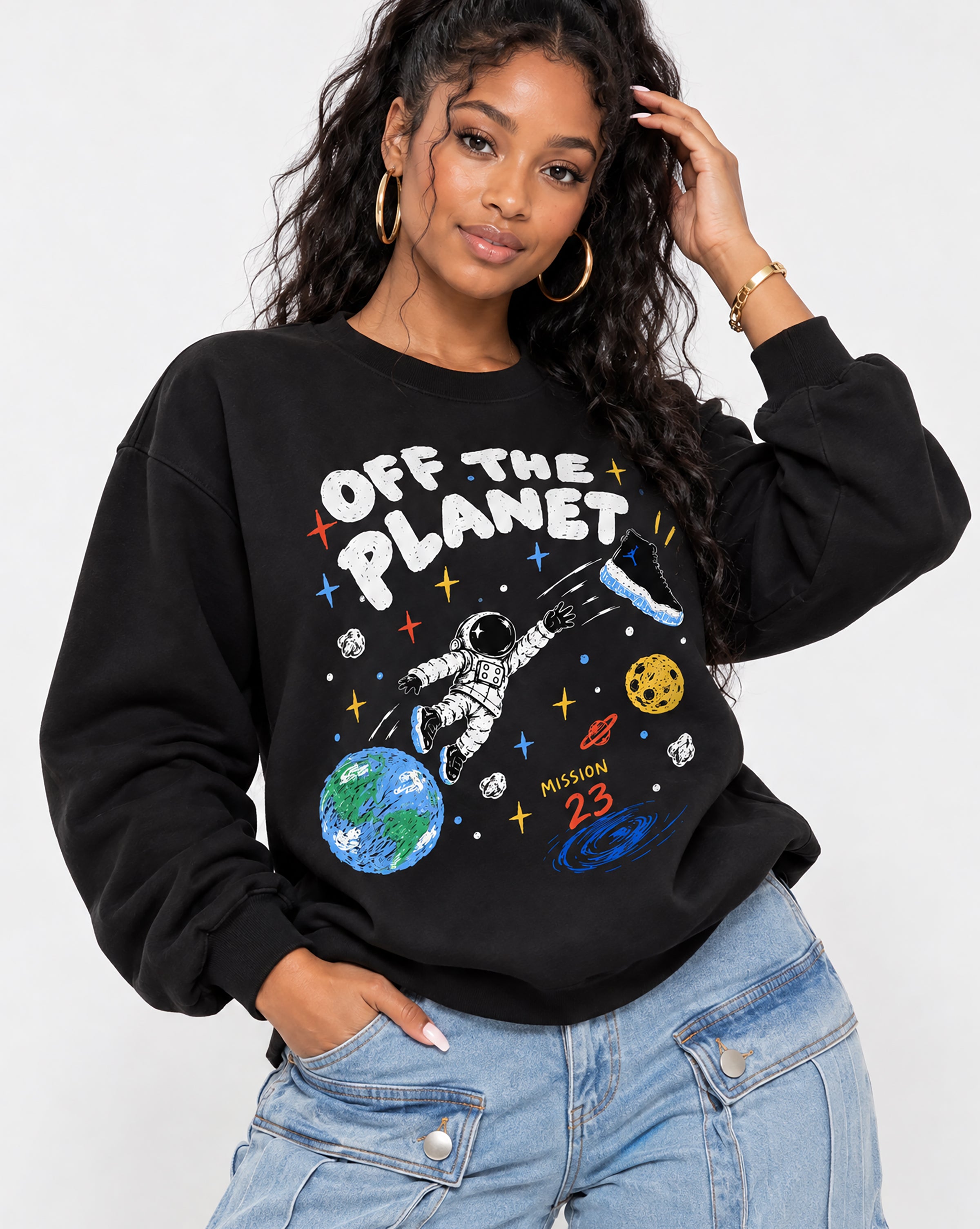 A woman wearing a black crewneck sweatshirt with a colorful space-themed graphic that reads 'OFF THE PLANET' and 'MILION 23', paired with light blue jeans, against a plain light gray background.