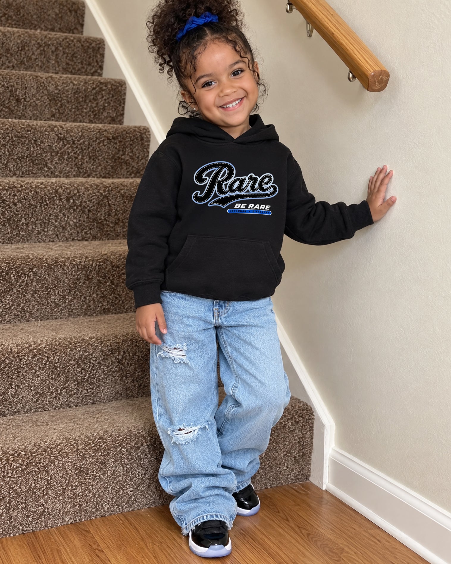 A young child wearing a black hoodie with 'Rare BE RARE' printed on the chest, paired with light blue distressed jeans and black sneakers, standing on carpeted stairs.