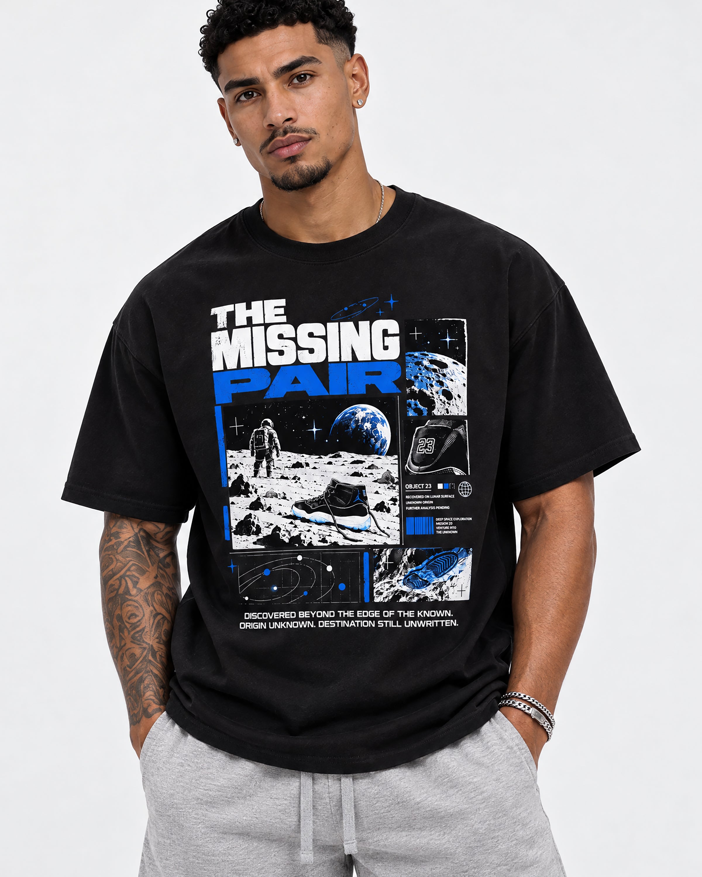 A man wearing a black graphic t-shirt with a space-themed print reading 'THE MISSING PAIR' and light gray sweatpants, standing against a light gray studio background.