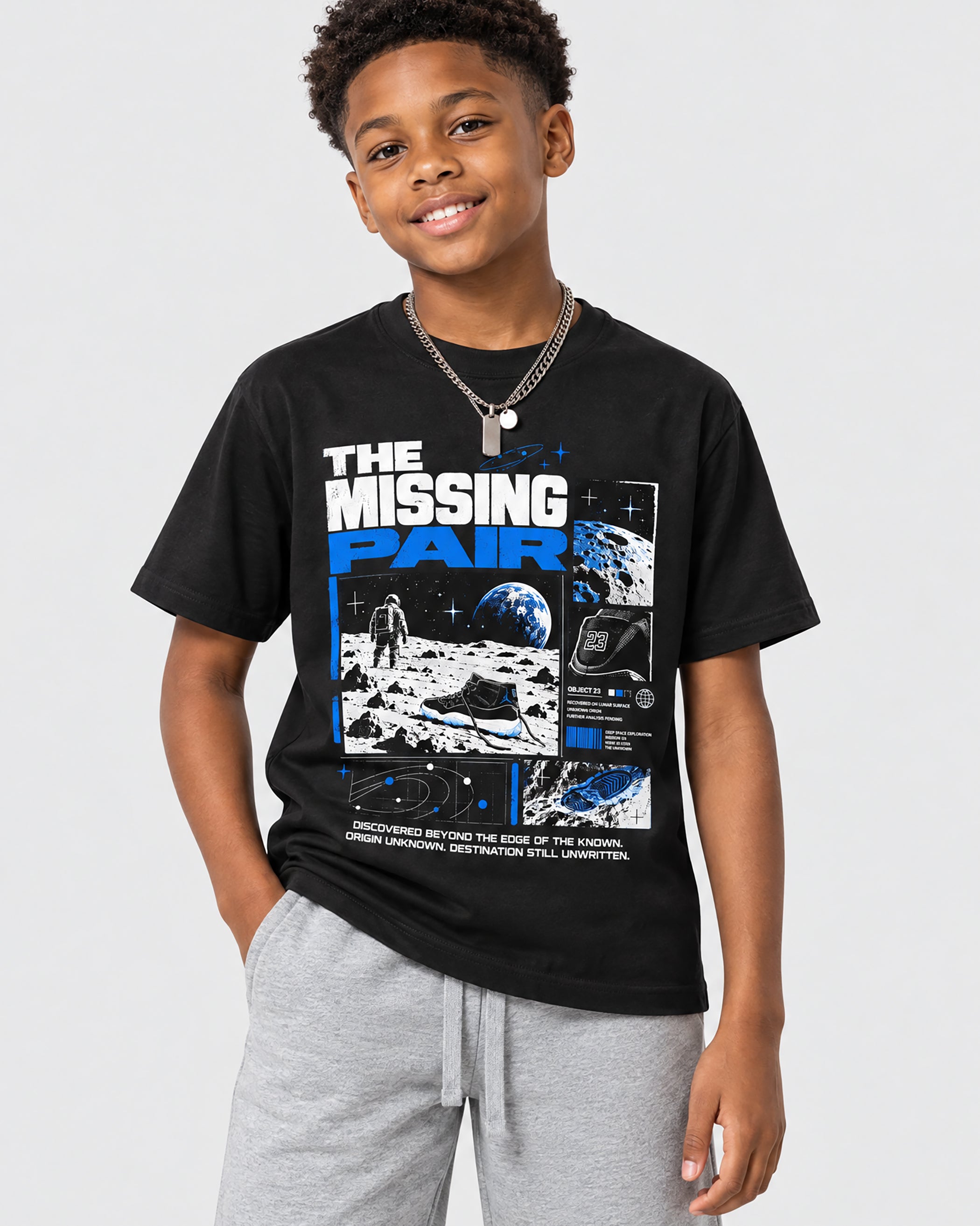 A young boy wearing a black graphic t-shirt with 'THE MISSING PAIR' text and space-themed illustrations, paired with gray sweatpants, against a light gray studio background.