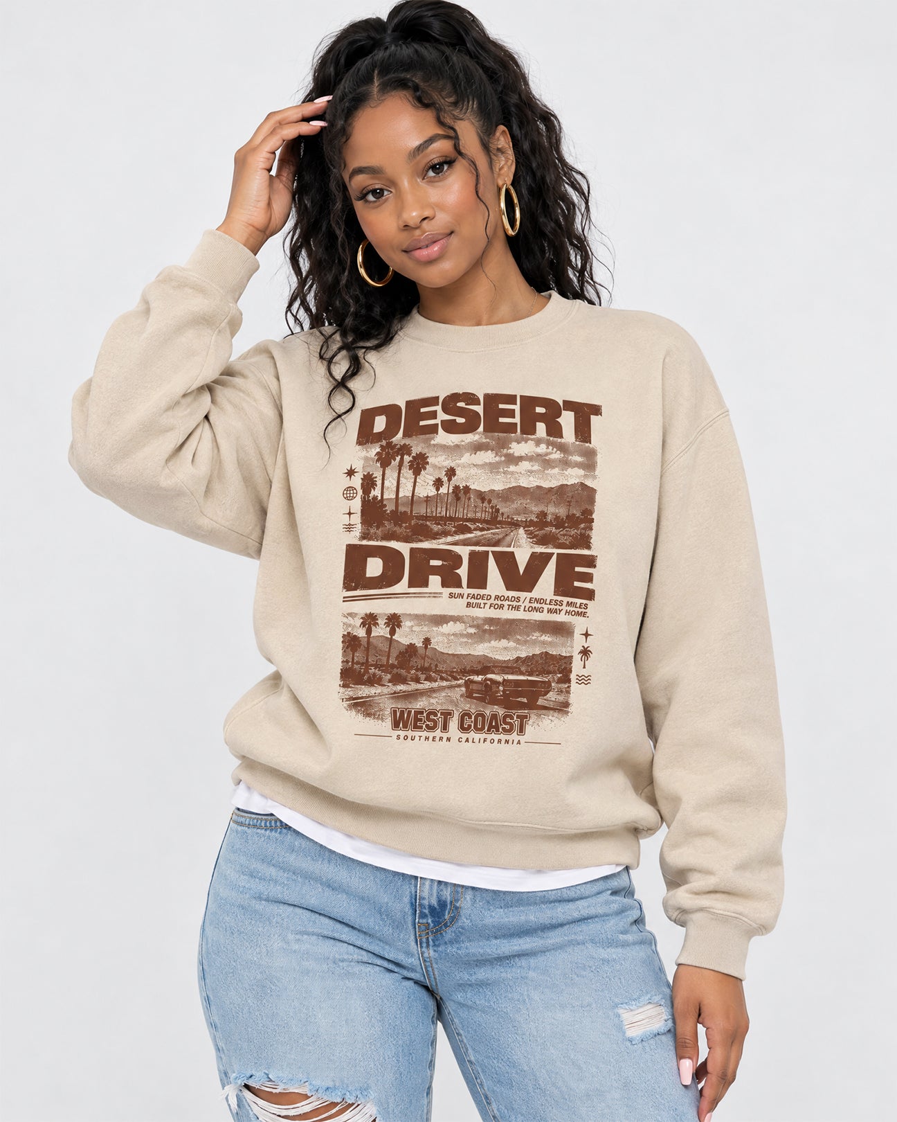 A woman modeling a beige crewneck sweatshirt with a brown retro graphic that reads 'DESERT DRIVE' and 'WEST COAST'. She is wearing light blue ripped jeans and gold hoop earrings against a plain light gray background.