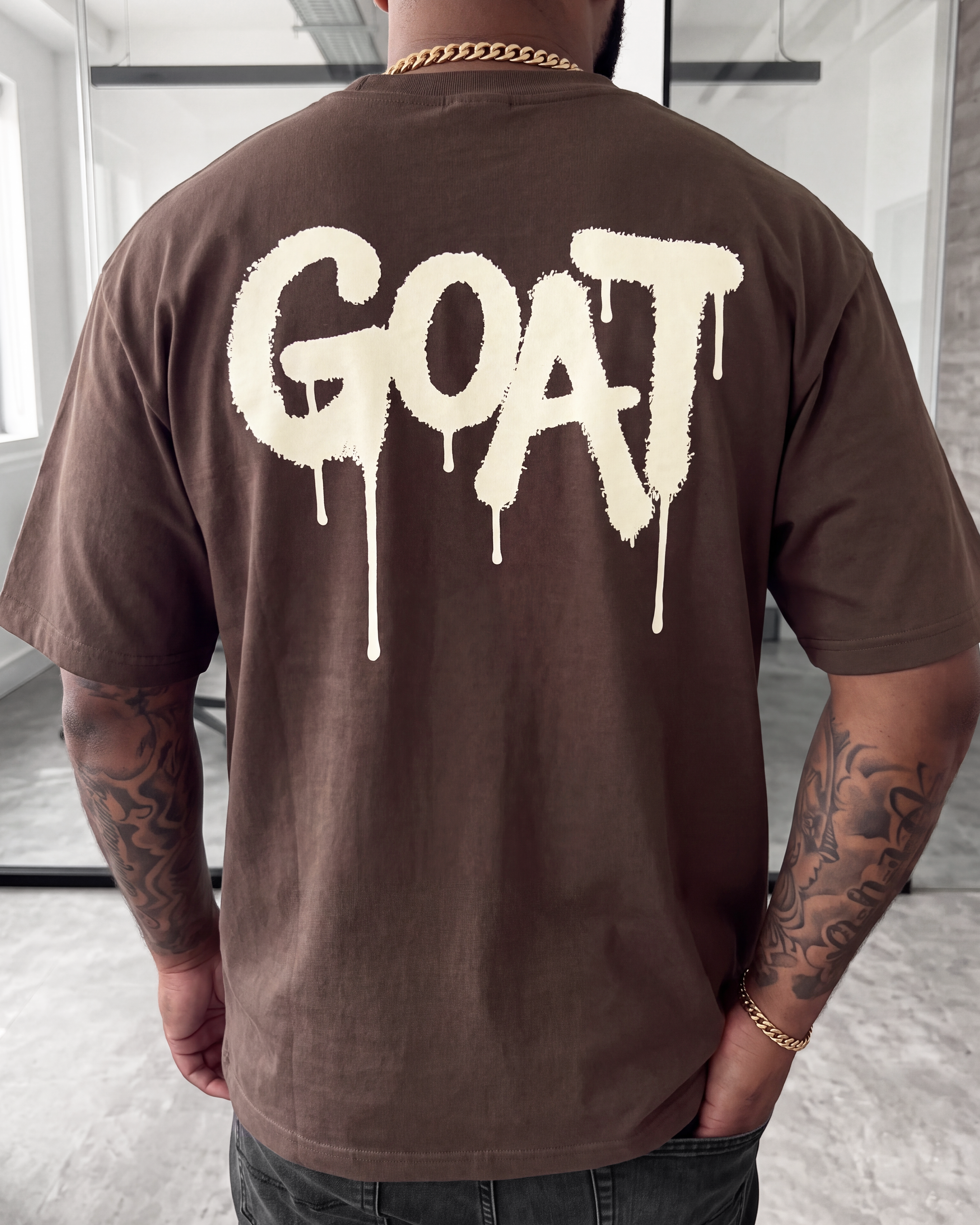 A man viewed from behind wearing a brown t-shirt with a large cream-colored dripping 'GOAT' logo on the back, set in a modern indoor space.