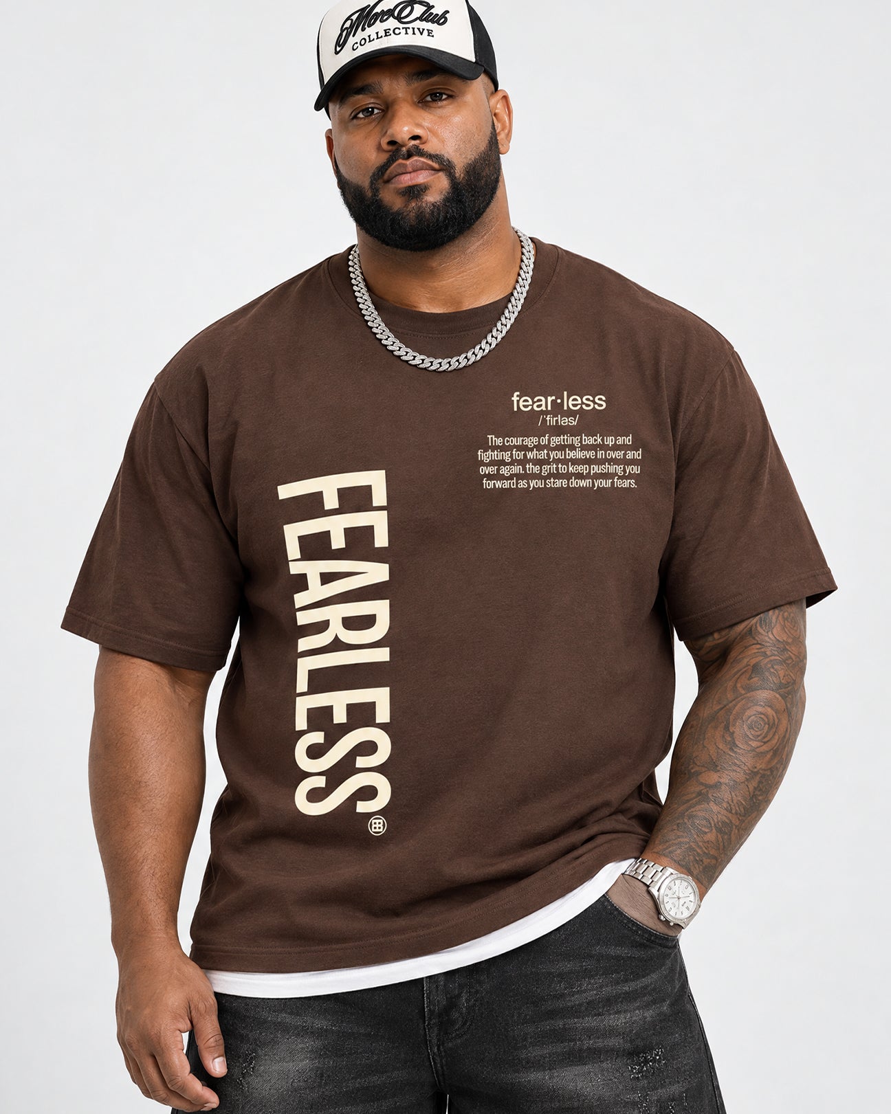 A man wearing a brown Fearless t-shirt with a large cream-colored vertical logo and a small quote on the chest, paired with a black and white cap, silver chain, and dark jeans.