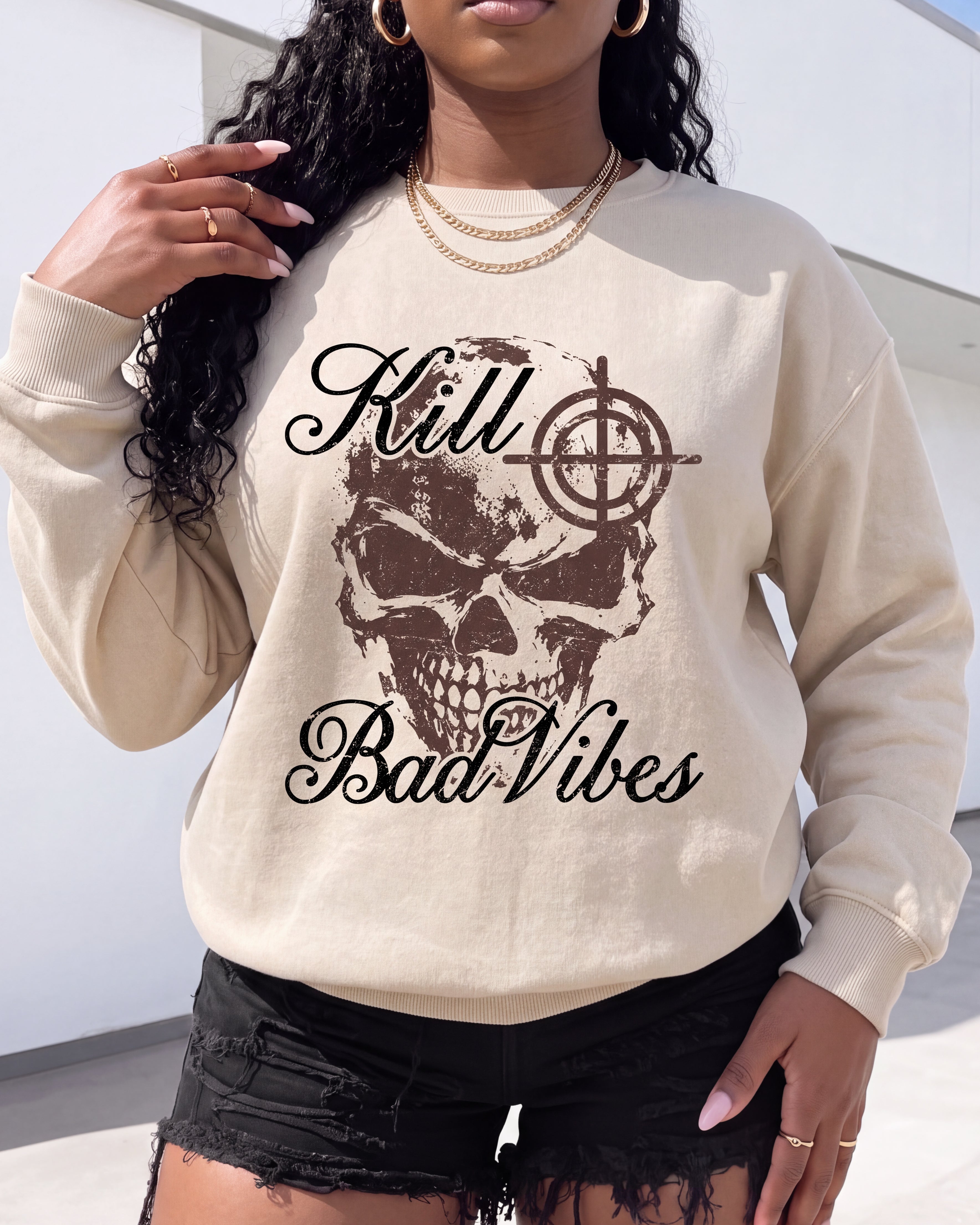 A woman wearing a cream-colored crewneck sweatshirt with a brown skull and target graphic and the text 'Kill Bad Vibes' printed on the front, paired with black distressed denim shorts.