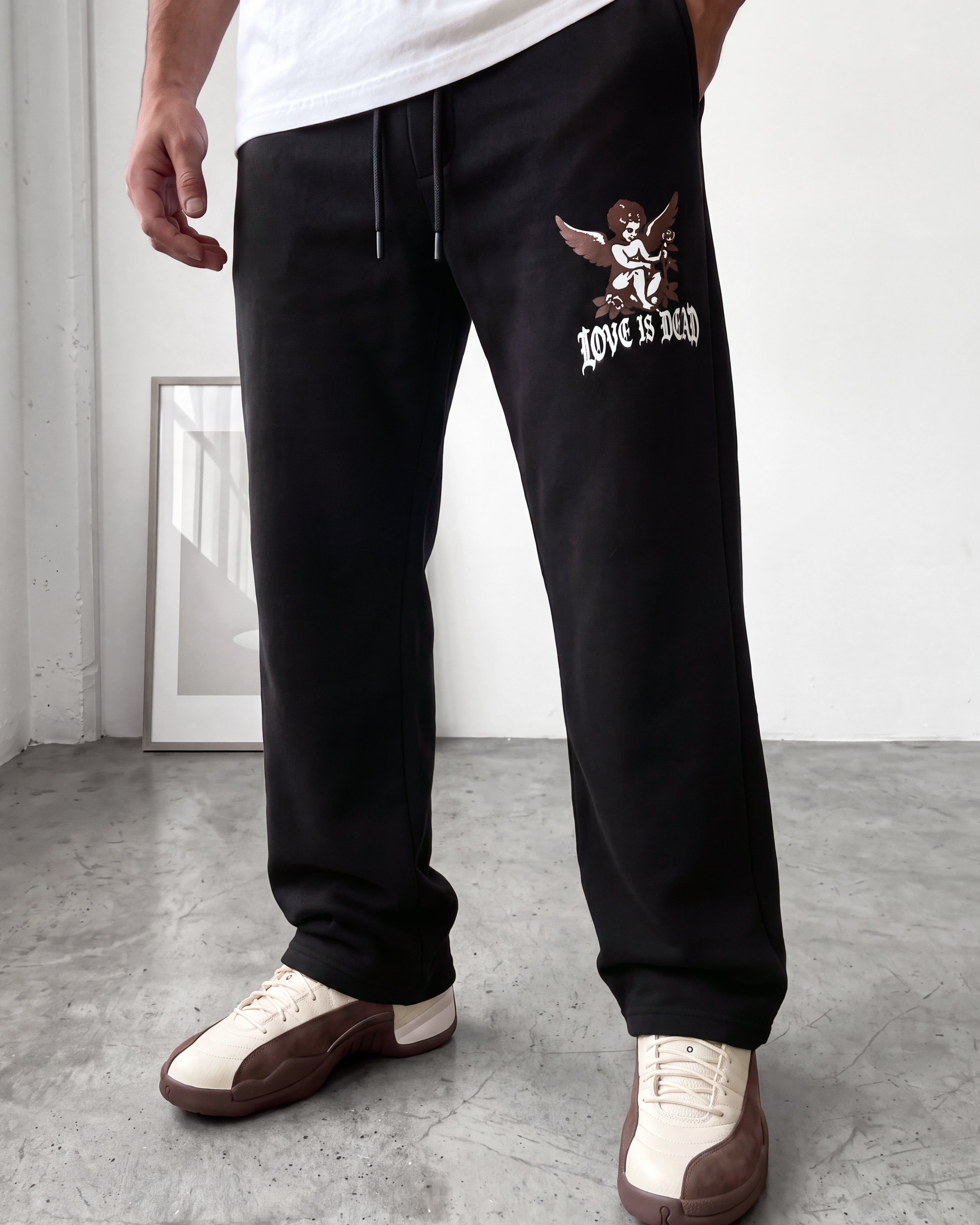 A person wearing black sweatpants with a graphic of an angel and the text 'LOVE IS DEAD' on the left thigh, paired with cream and brown sneakers in a minimalist indoor setting.