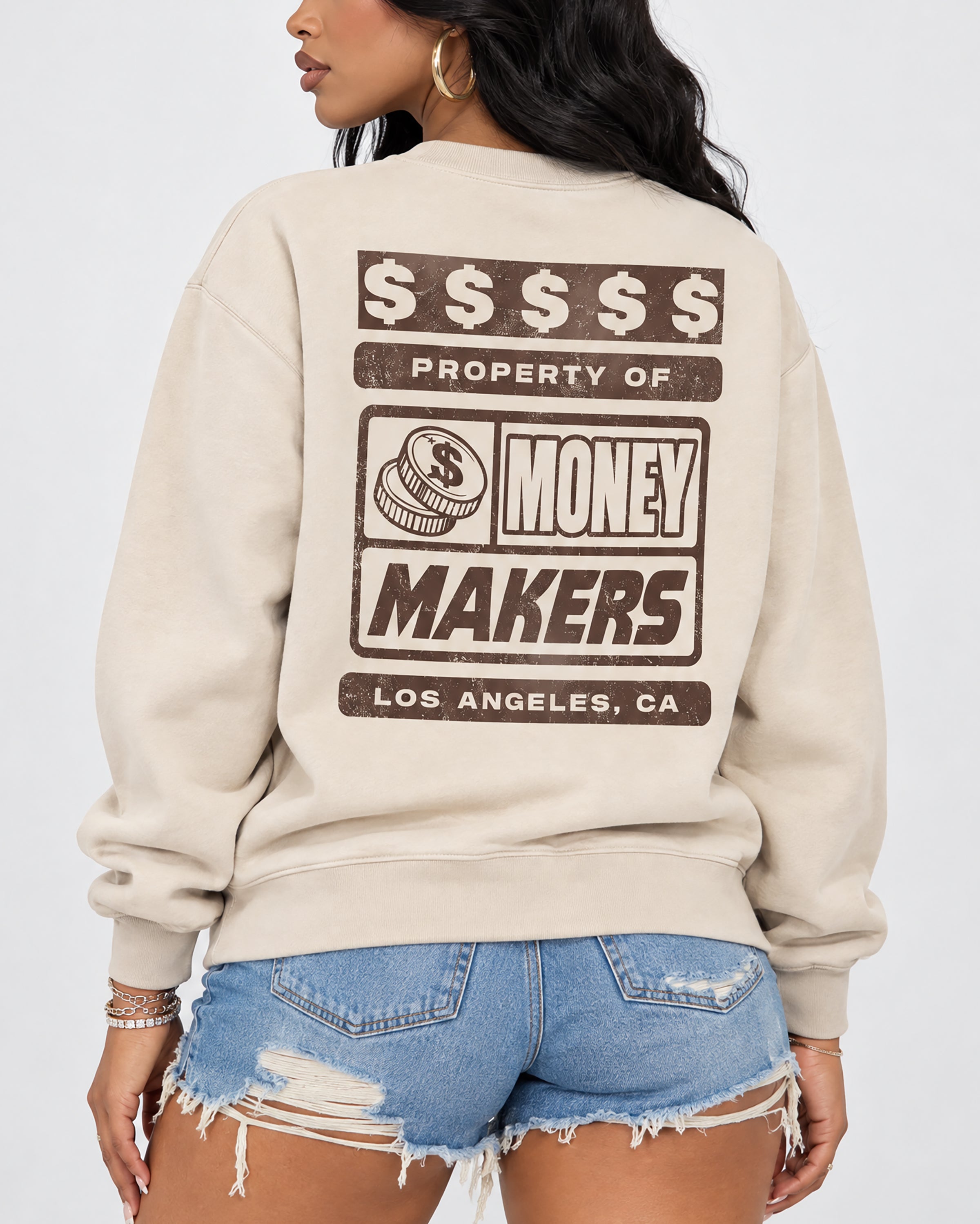 A woman shown from the back wearing a cream-colored crewneck sweatshirt with a large brown graphic print that reads '$$$$$$ PROPERTY OF MONEY MAKERS LOS ANGELES, CA' above distressed denim shorts.