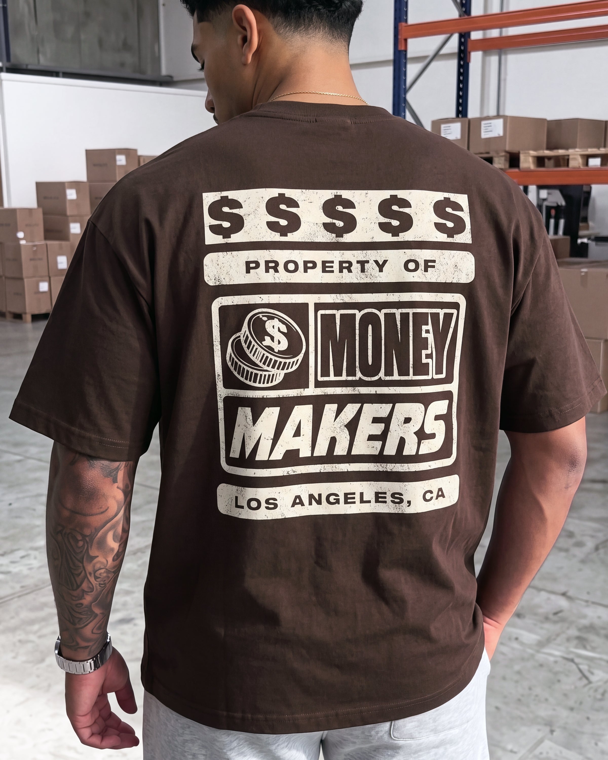 A man viewed from behind wearing a brown t-shirt with a large graphic print on the back that reads 'PROPERTY OF MONEY MAKERS LOS ANGELES, CA' with a coin icon, in a warehouse setting.