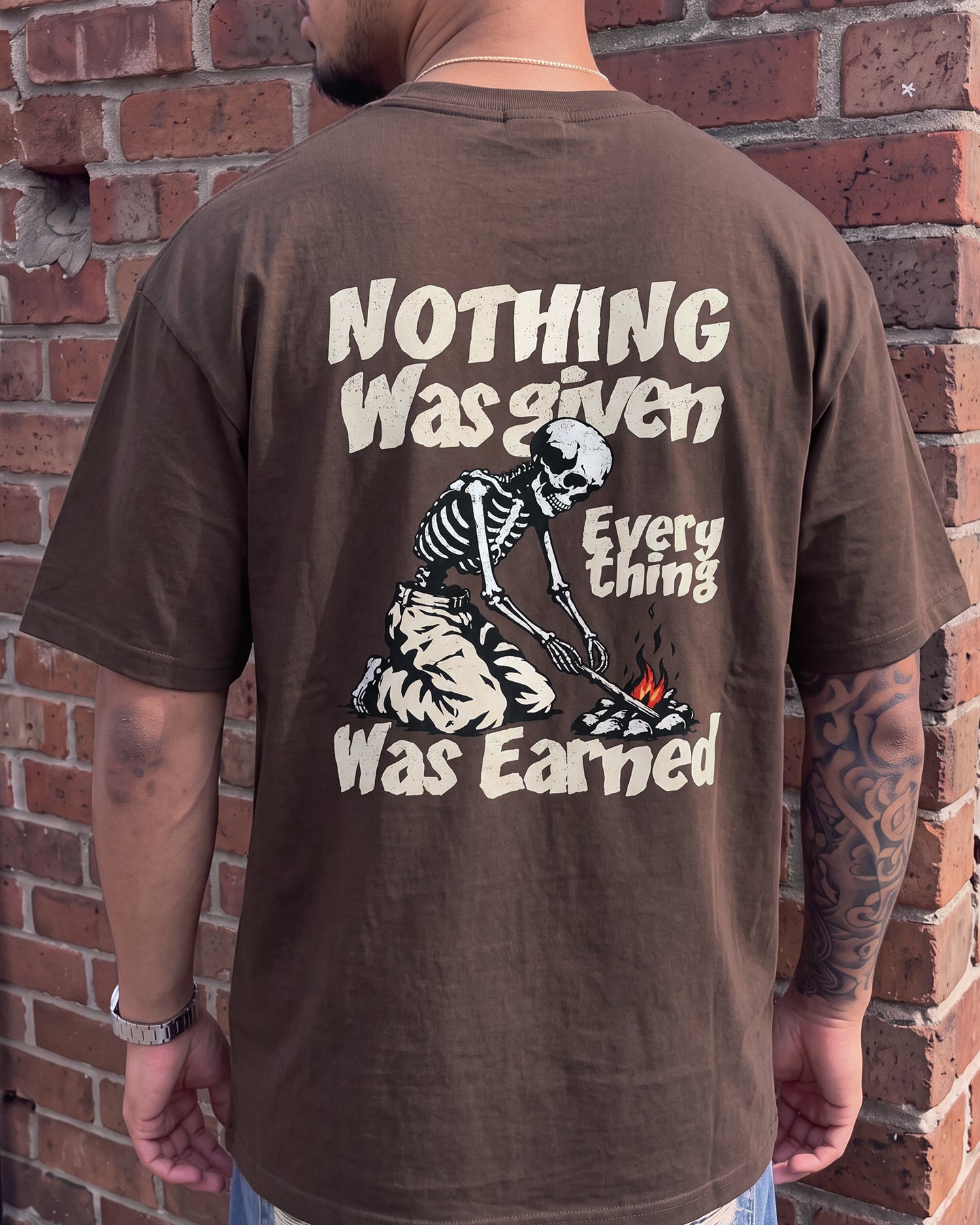 A man viewed from behind wearing a brown graphic t-shirt with a skeleton and text design, standing against a red brick wall.