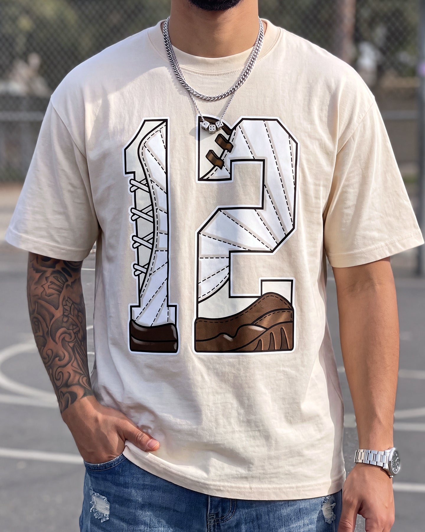 A man wearing a cream-colored t-shirt with a large graphic of the number 12 filled with shoe illustrations in brown and white, paired with blue jeans.