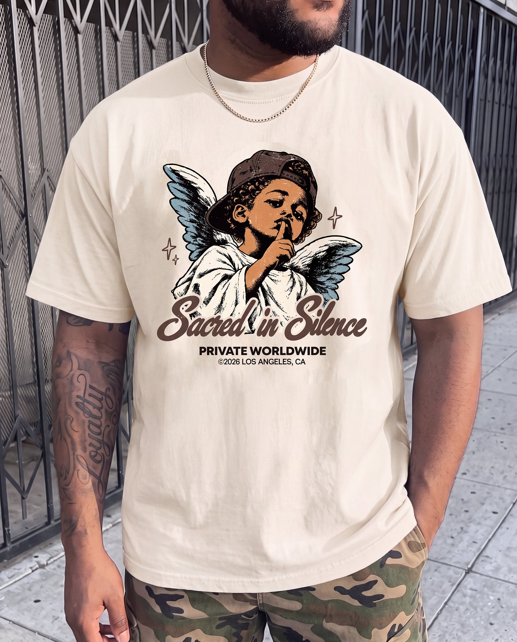 A man wearing a cream-colored graphic t-shirt with a vintage-style angel illustration and the text 'Sacred in Silence' and 'PRIVATE WORLDWIDE LOS ANGELES, CA'.