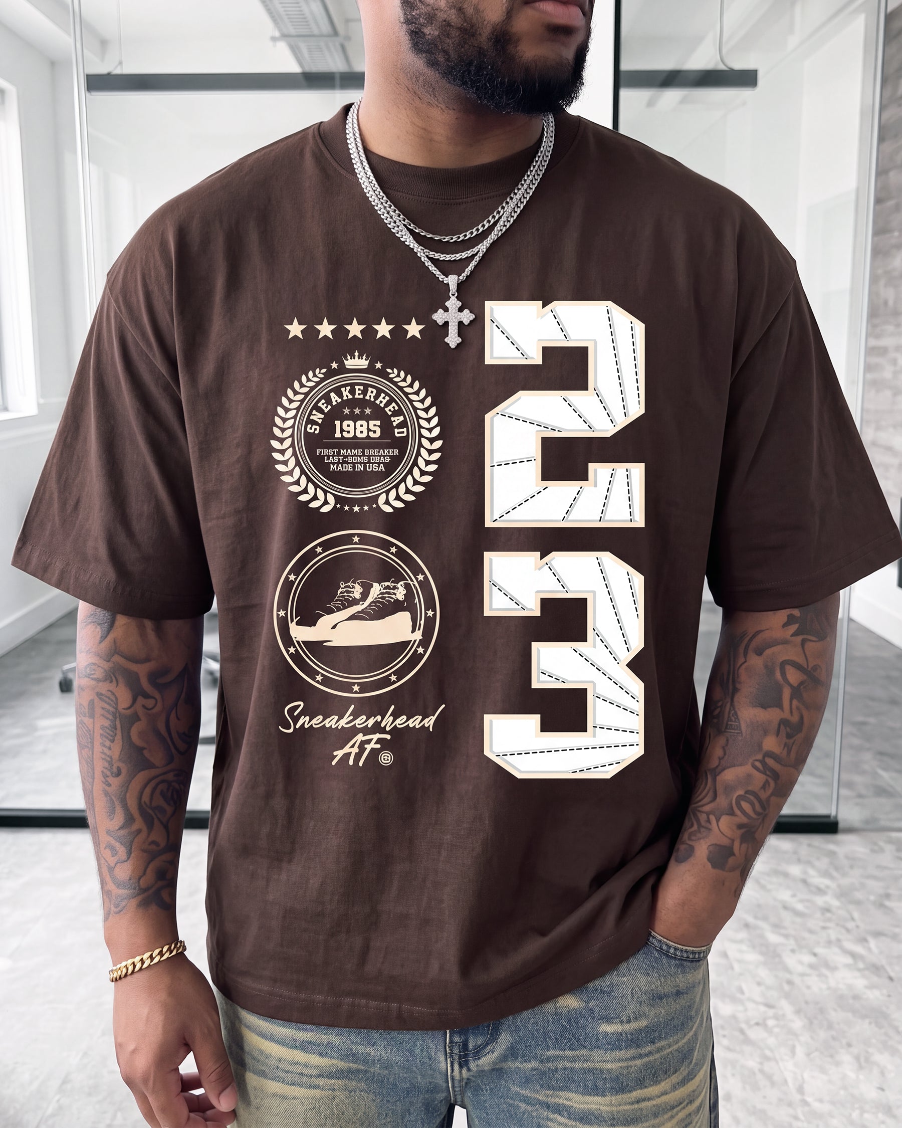 A man wearing a brown short-sleeve graphic t-shirt with a large white '23' print and circular 'Basketball 1985 Sneakerhead AF' graphics on the front, paired with distressed blue jeans and silver jewelry.