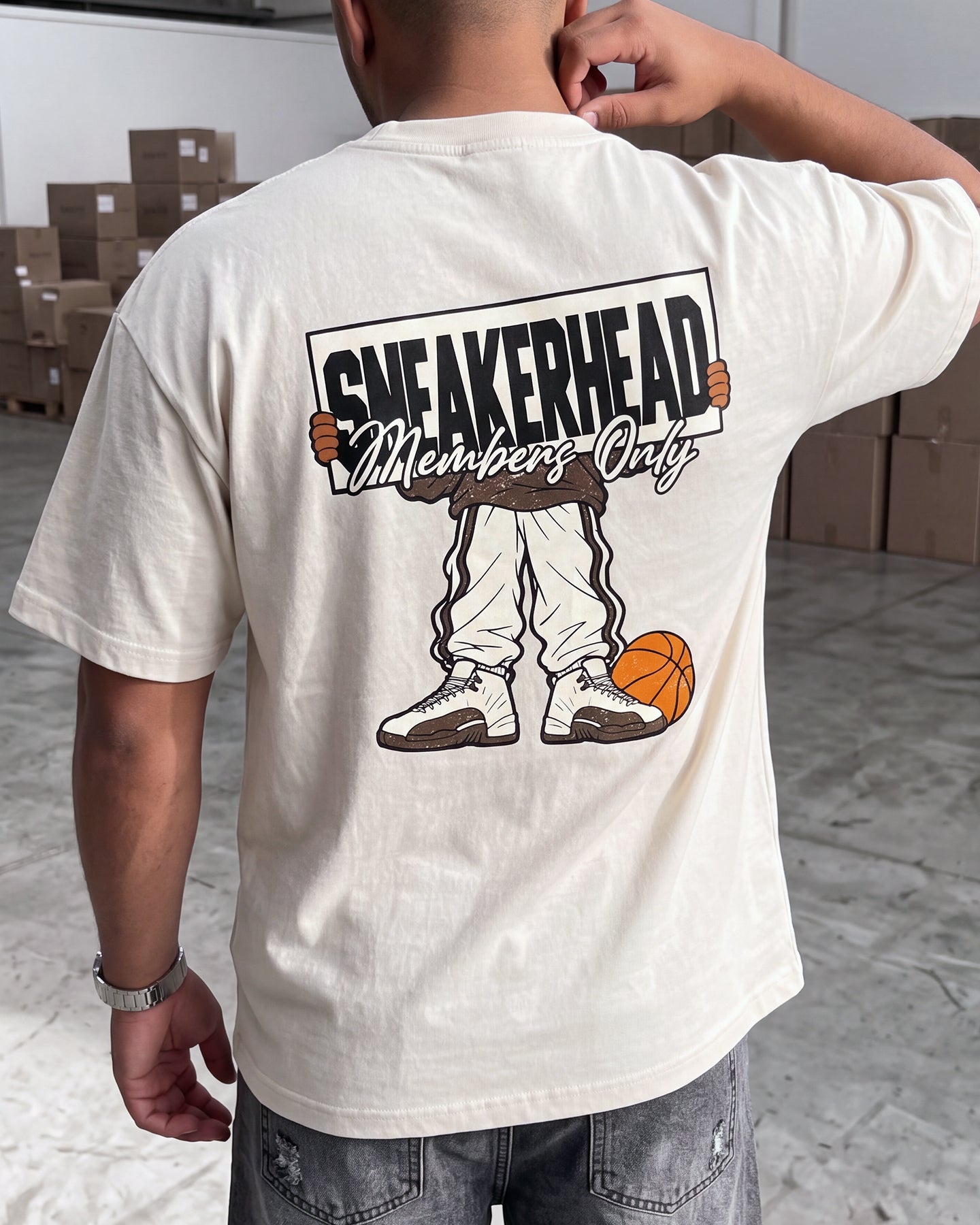A man viewed from behind wearing a cream-colored t-shirt with a large graphic on the back that reads 'SNEAKERHEAD Members Only' above a cartoon figure in sneakers and a basketball.