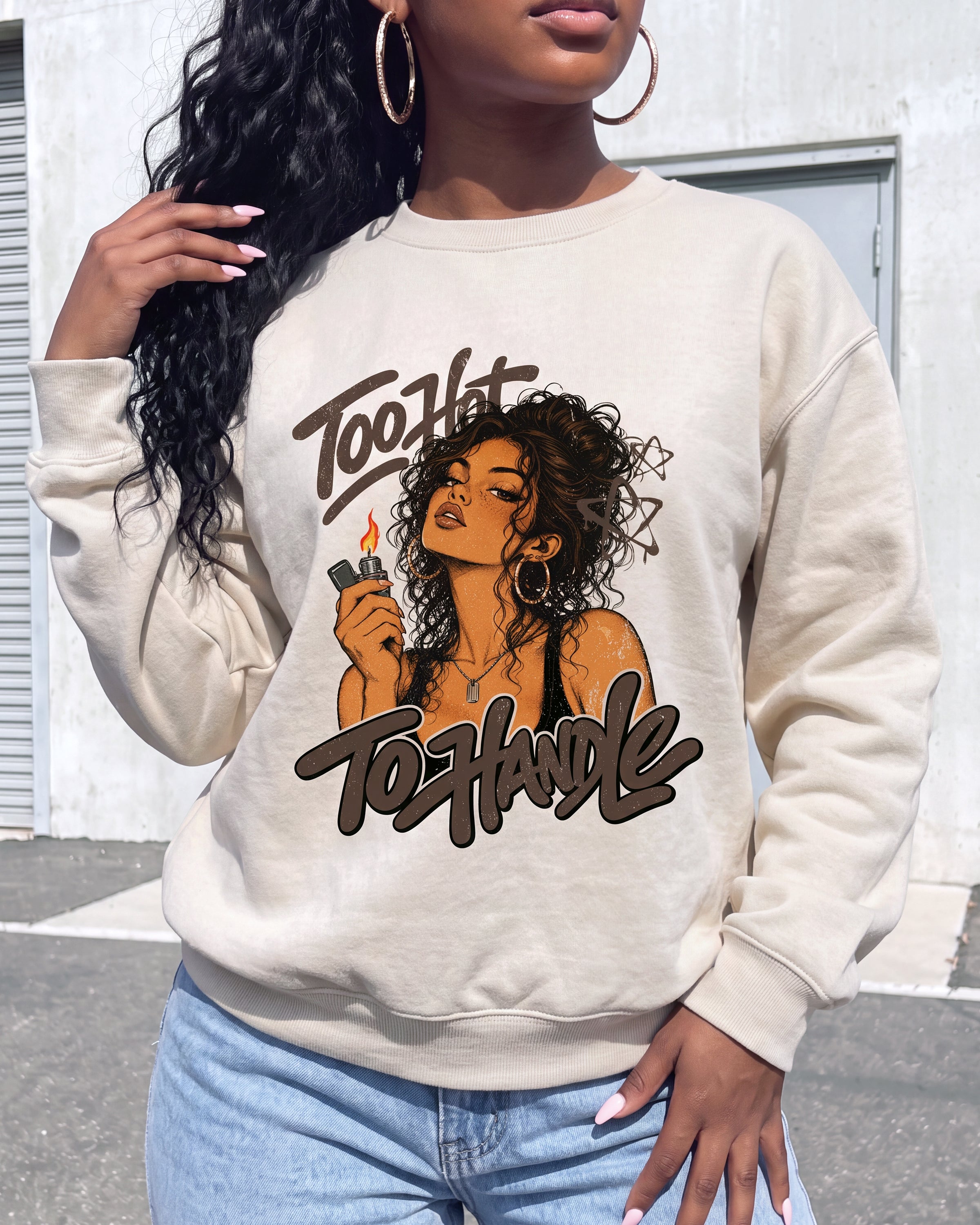 A woman wearing a beige crewneck sweatshirt with a graphic print that reads 'Too Hot To Handle' and shows a woman with curly hair holding a lighter. She is wearing light blue jeans and large gold hoop earrings, standing outdoors against a light gray wall.