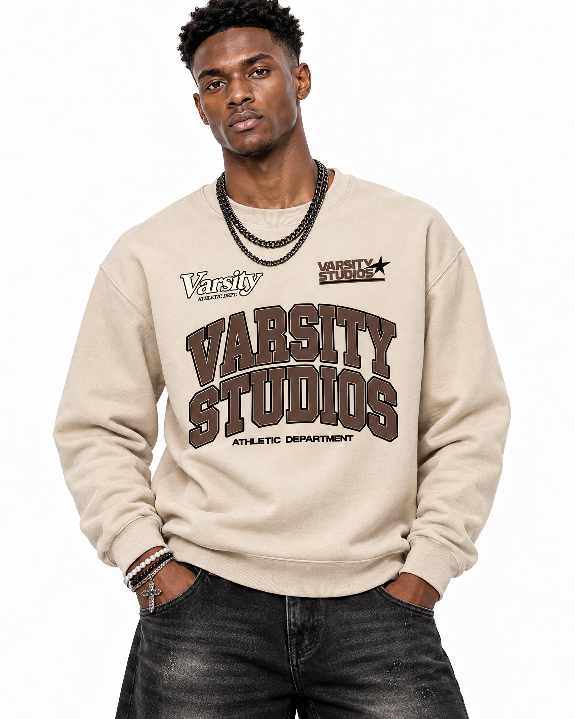 A man wearing a beige crewneck sweatshirt with 'Varsity Studios Athletic Department' printed in brown on the front, paired with dark gray jeans and layered black chain necklaces, against a plain white background.