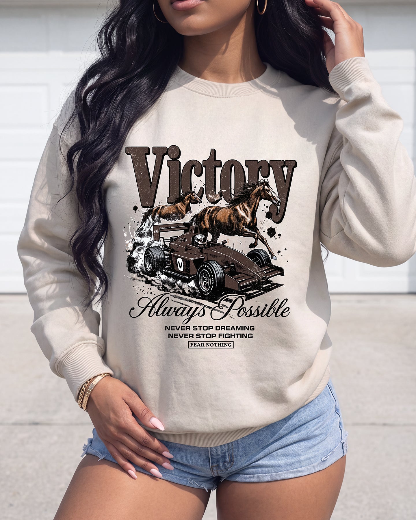 A woman wearing a beige crewneck sweatshirt with a graphic print of a racehorse on a racecar and the words 'Victory' and 'Always Possible'.