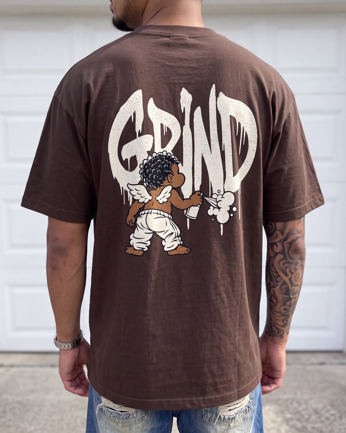 A man viewed from behind wearing a brown t-shirt with a large white 'GRIND' graphic and cartoon character print on the back, paired with distressed blue jeans in front of a white garage door.