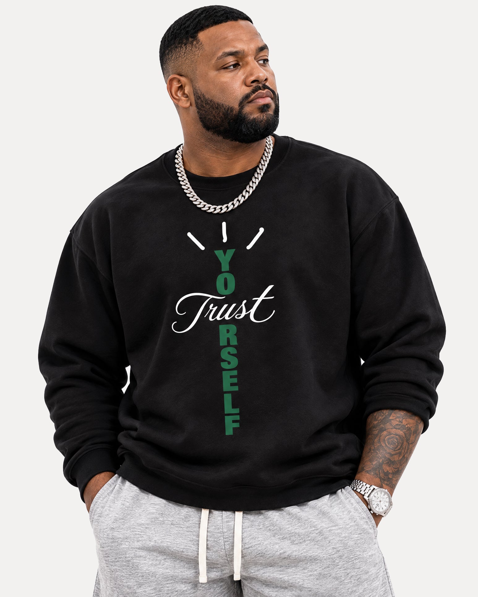 A man wearing a black crewneck sweatshirt with a graphic print that reads 'Trust Yourself' in green and white, paired with gray sweatpants and a silver chain necklace, against a light gray background.