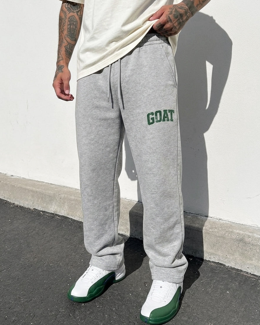 A person wearing heather gray sweatpants with green 'GOAT' text on the left thigh, paired with a cream t-shirt and green-and-white sneakers, standing against a white wall.