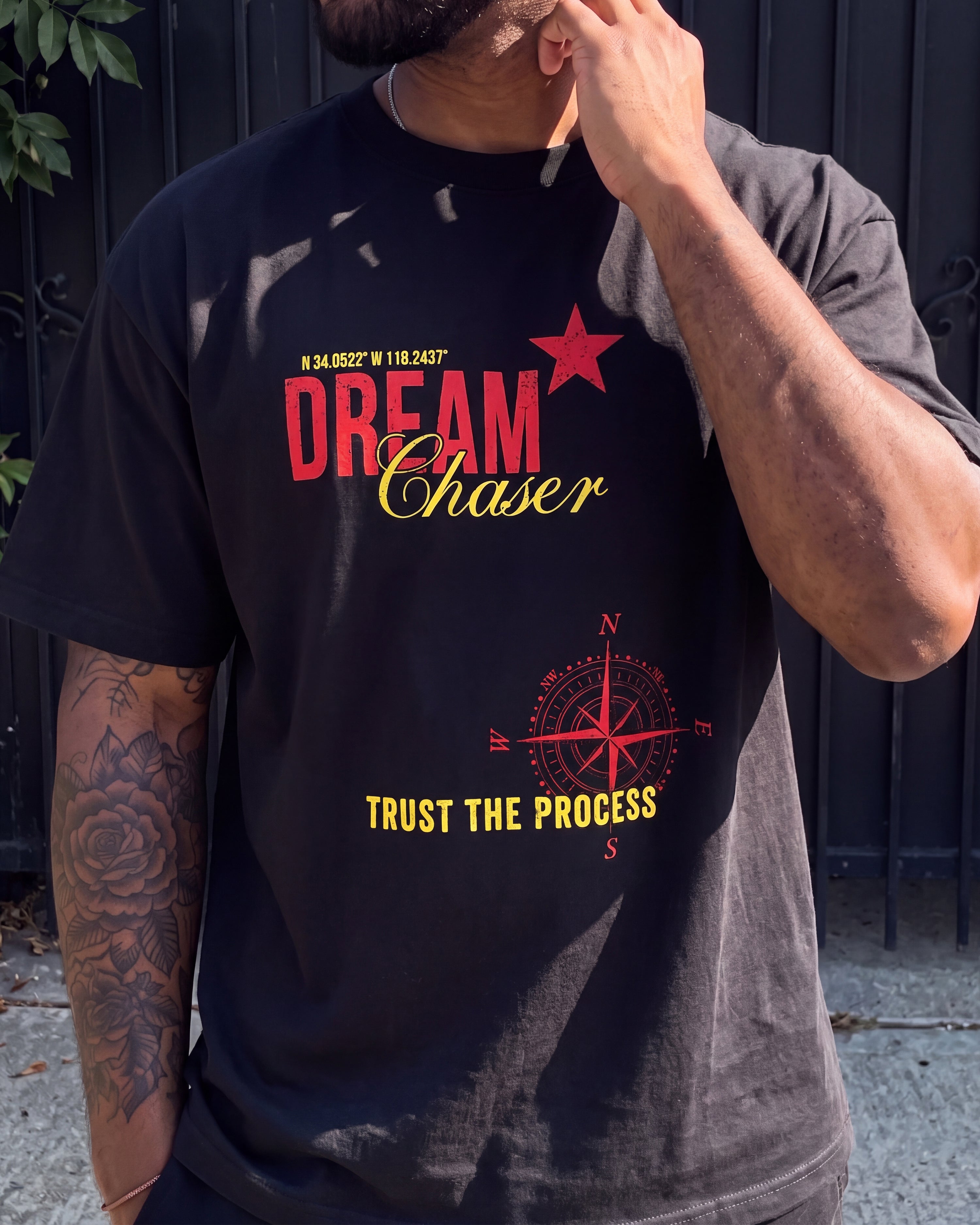 A man wearing a black graphic t-shirt with red and yellow text reading 'DREAM Chaser' and 'TRUST THE PROCESS' along with a red star and compass design, set against a dark fence background.
