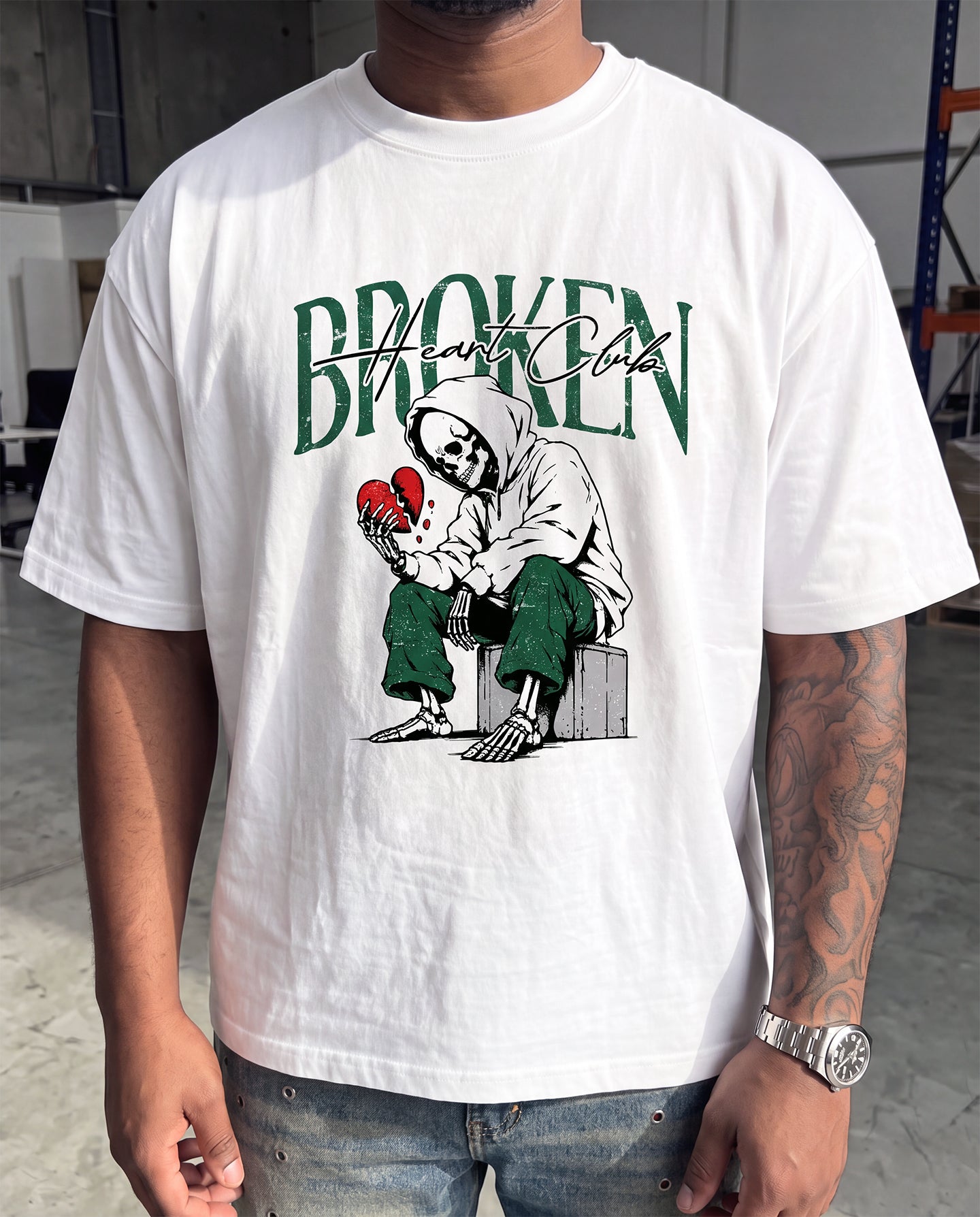 A man wearing a white graphic t-shirt with a green and black 'Broken Heart Club' design, paired with blue jeans in an industrial warehouse setting.
