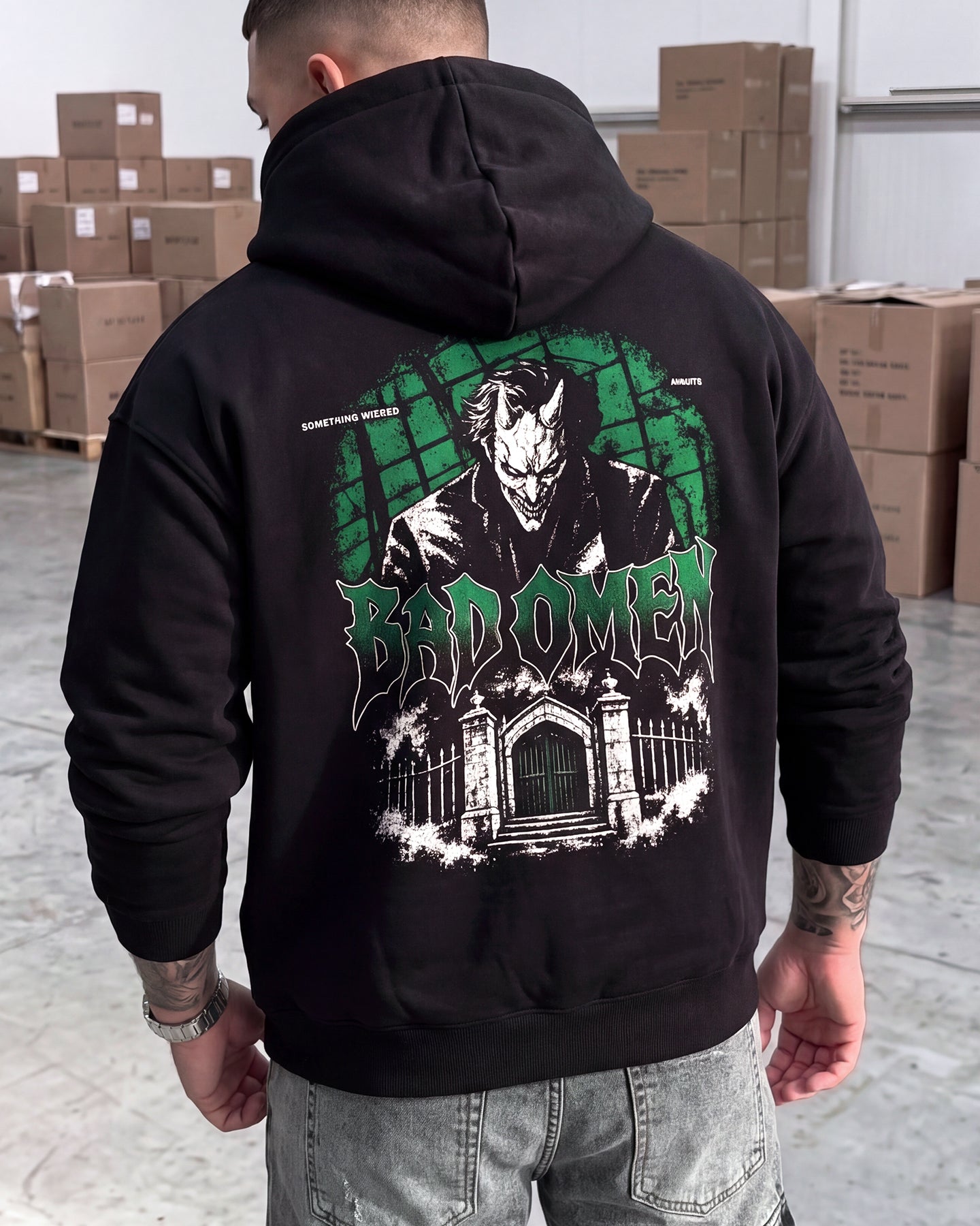 A man viewed from behind wearing a black hoodie with a large green and white graphic print on the back. The graphic features the text 'BADOMEN' and a gothic illustration.