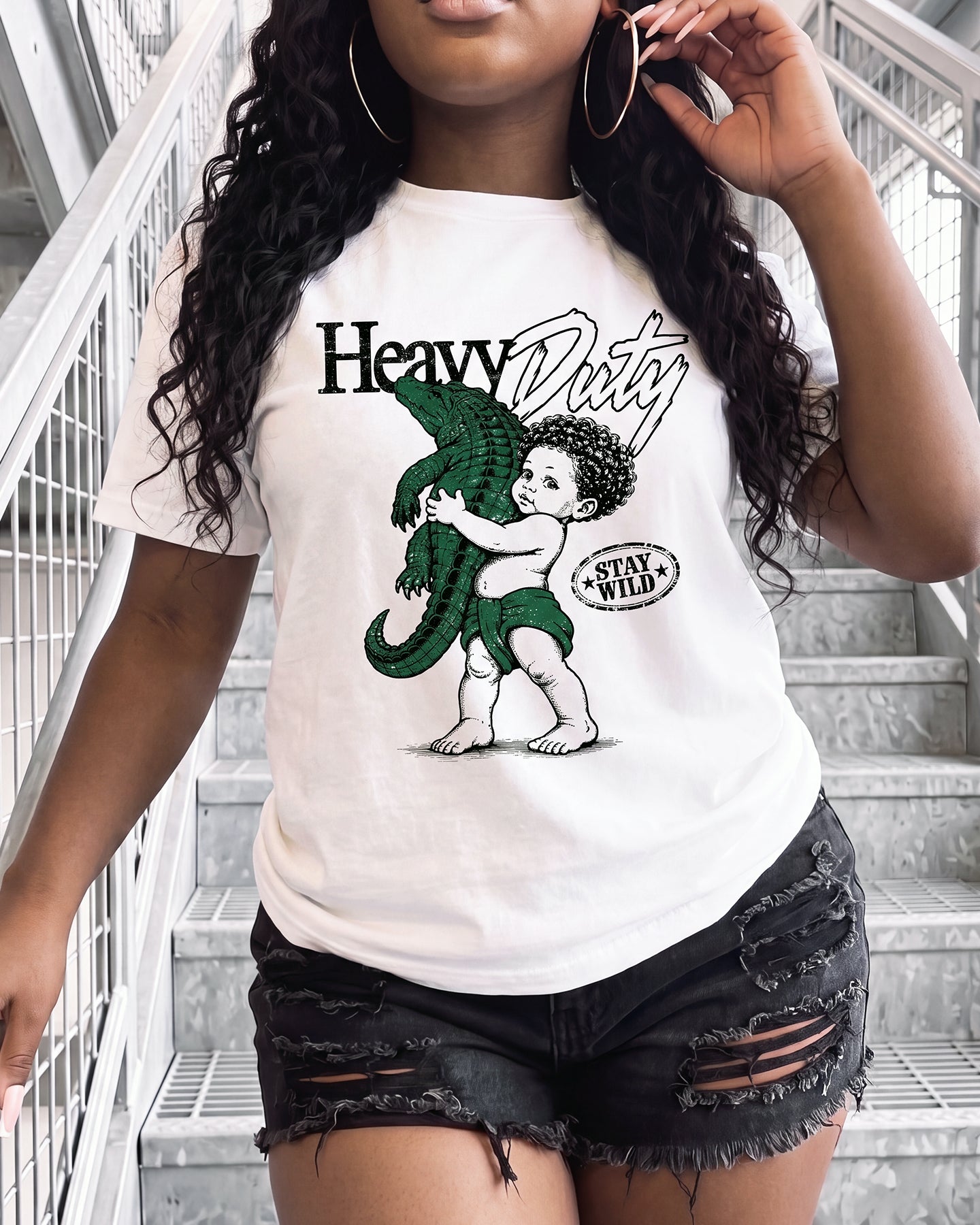 A woman wearing a white graphic t-shirt with a green and black print of a child hugging an alligator, paired with distressed black denim shorts, standing on outdoor concrete stairs.