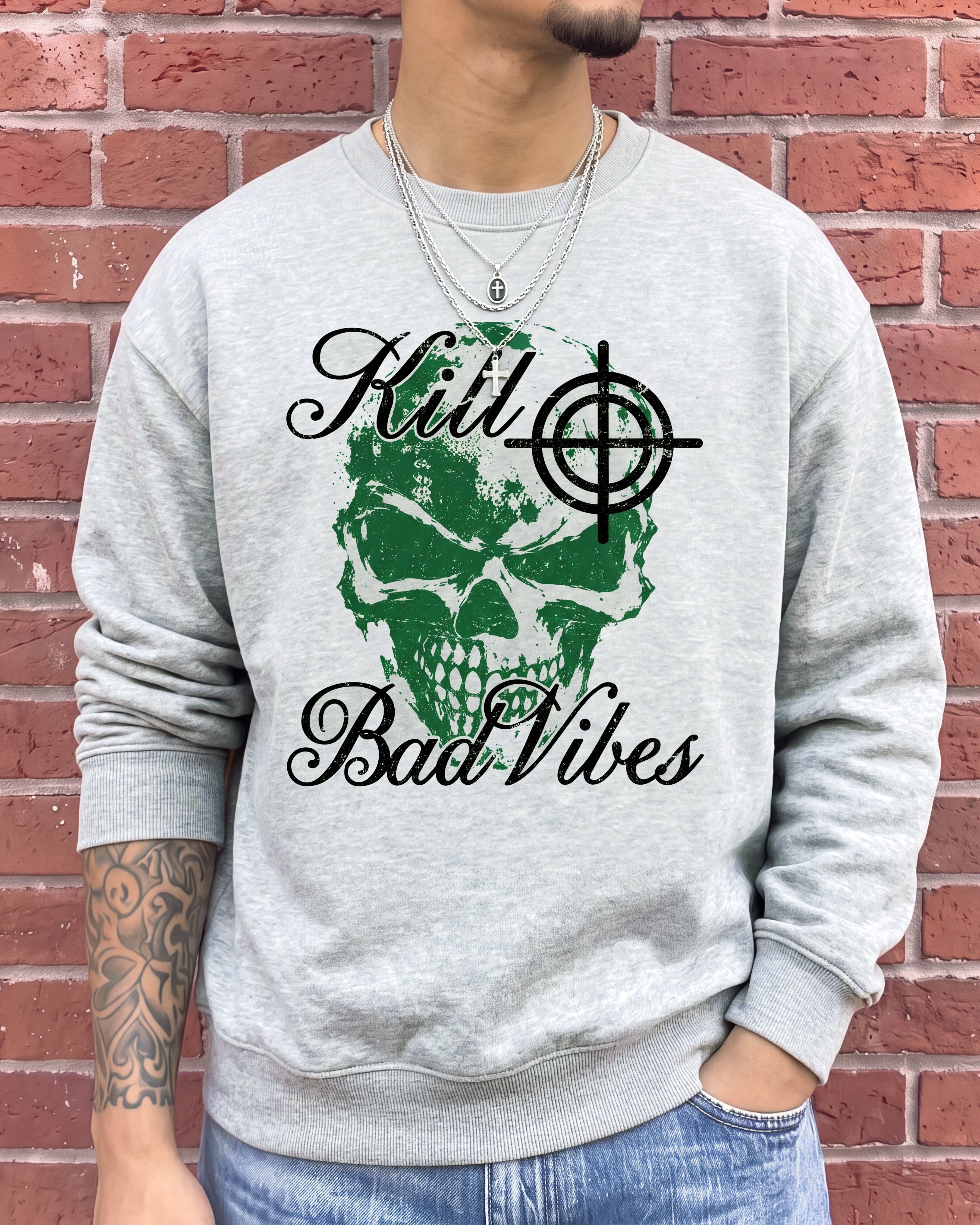 A man wearing a light gray crewneck sweatshirt with a green skull and black 'Kill Bad Vibes' graphic, standing against a red brick wall.