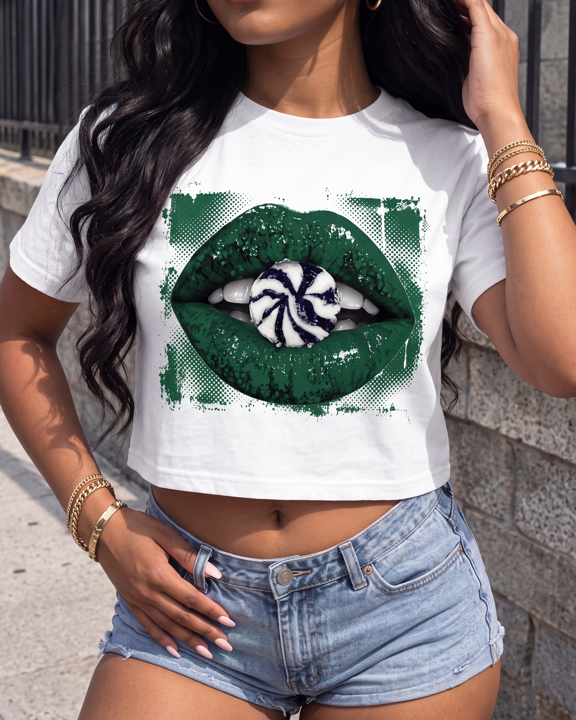 A woman wearing a white crop top t-shirt with a green graphic of lips holding a peppermint candy, paired with denim shorts.