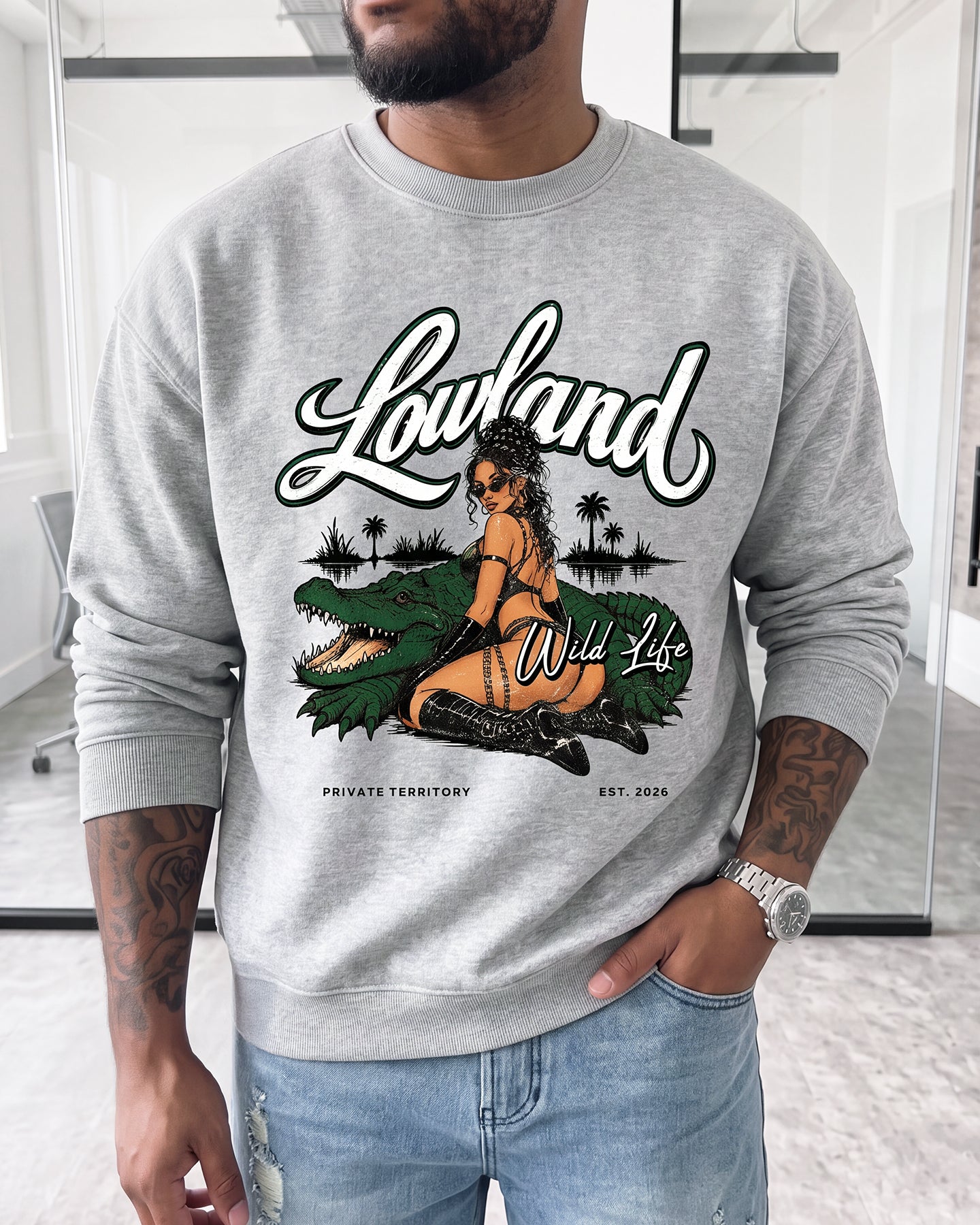 A man wearing a light gray crewneck sweatshirt with a graphic print of a woman sitting on a green alligator, featuring the text 'Lowland Wild Life' and 'PRIVATE TERRITORY'.