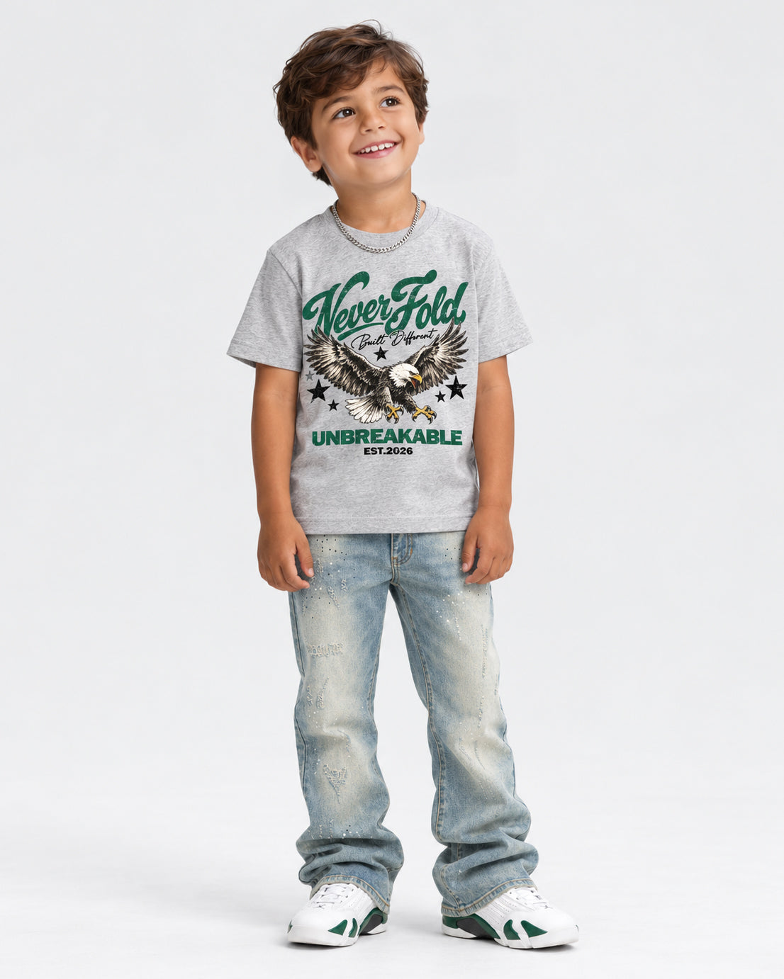 A young boy smiling and wearing a light gray graphic t-shirt with a green eagle design and the words 'Never Fold' and 'UNBREAKABLE', paired with light-wash jeans and white sneakers, standing against a light gray studio background.