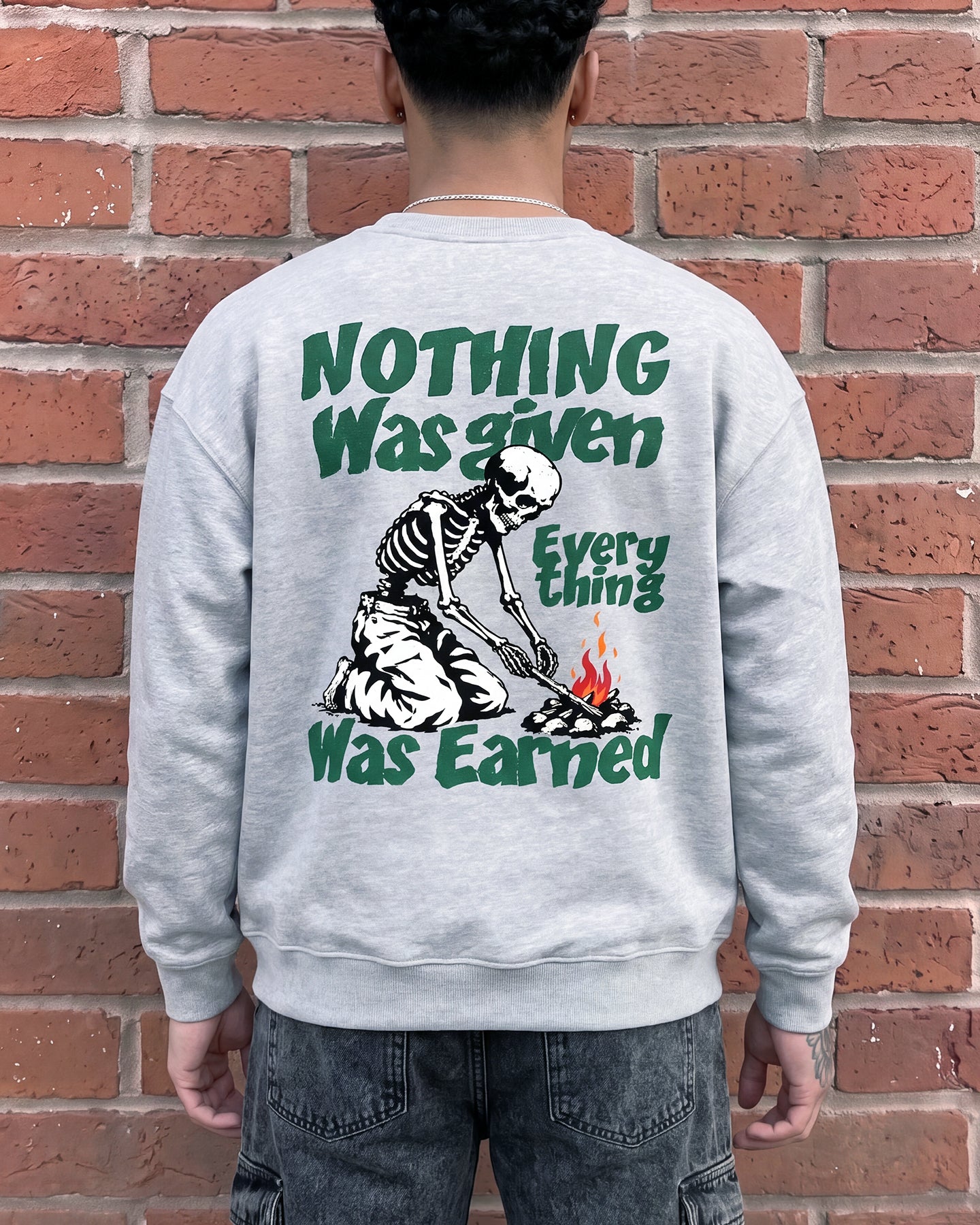 A person viewed from behind wearing a light gray crewneck sweatshirt with a large graphic print on the back. The print features a skeleton sitting by a campfire with the text 'NOTHING Was given Everything Was Earned' in dark green.