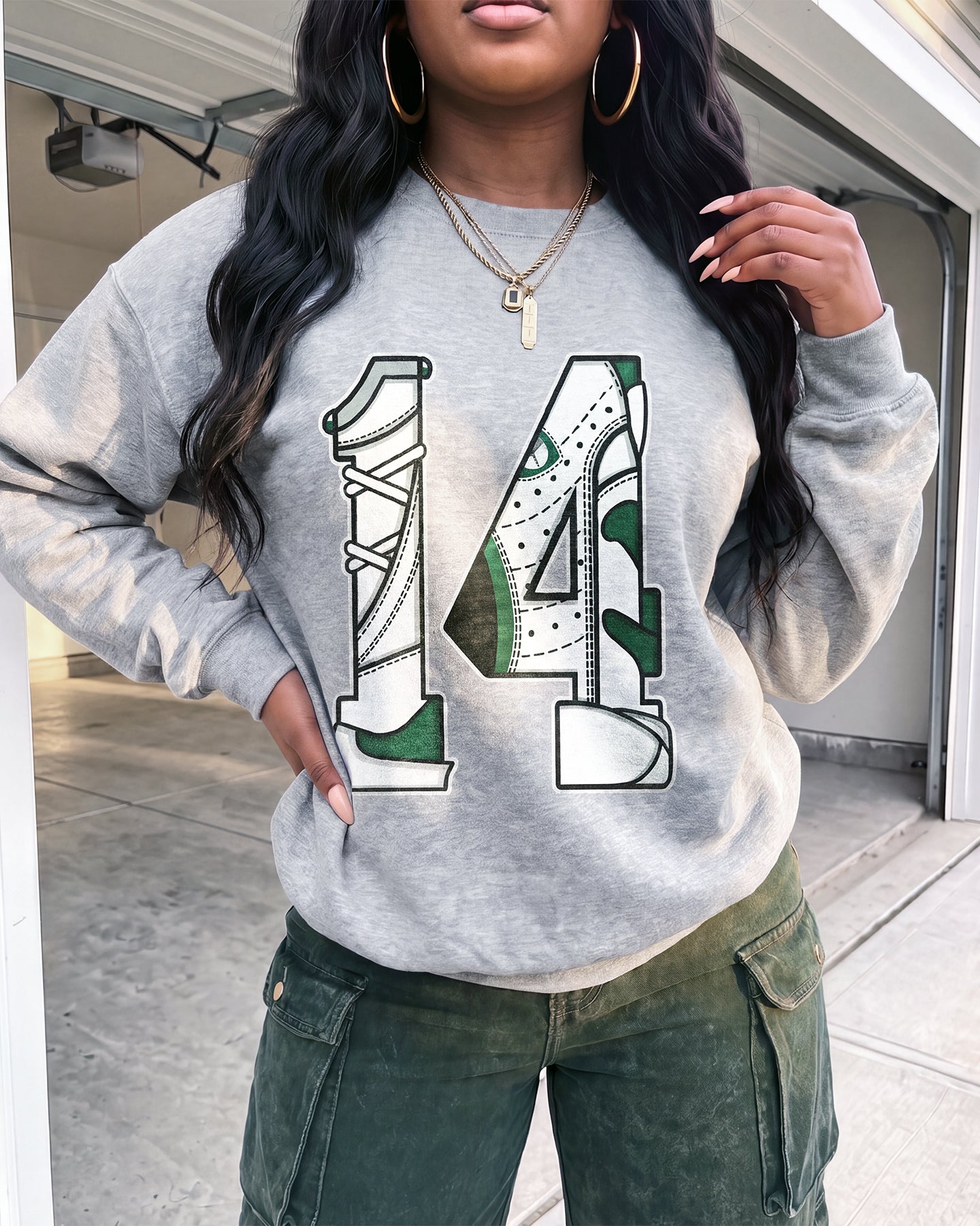 A woman wearing a heather gray crewneck sweatshirt with a graphic of two high-top sneakers forming the number 14, paired with dark green cargo pants.