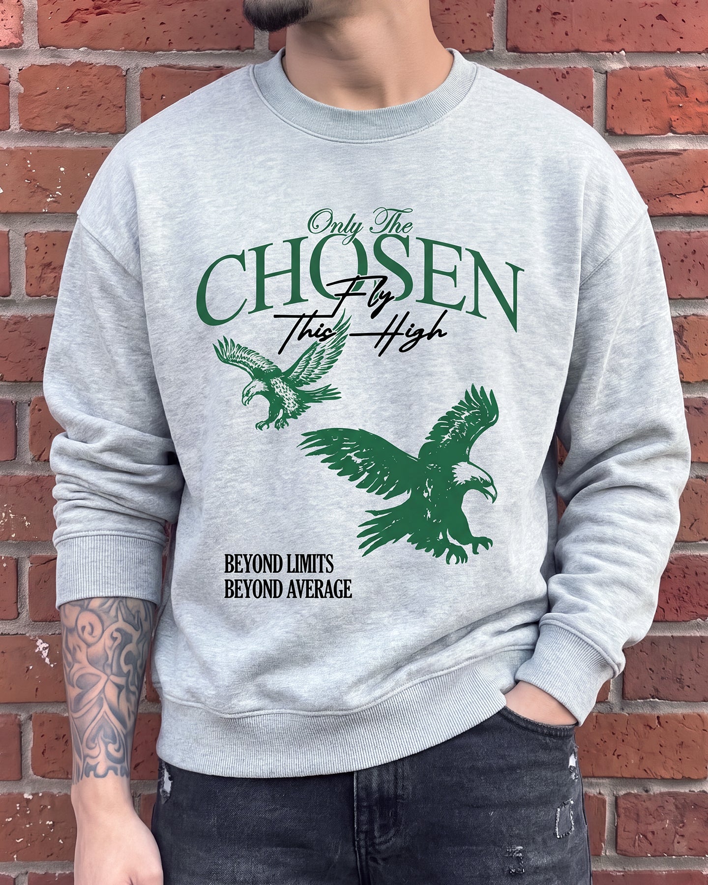 A man wearing a light gray crewneck sweatshirt with a green eagle graphic and the text 'Only The CHOSEN By This High' and 'BEYOND LIMITS BEYOND AVERAGE.' standing against a red brick wall.