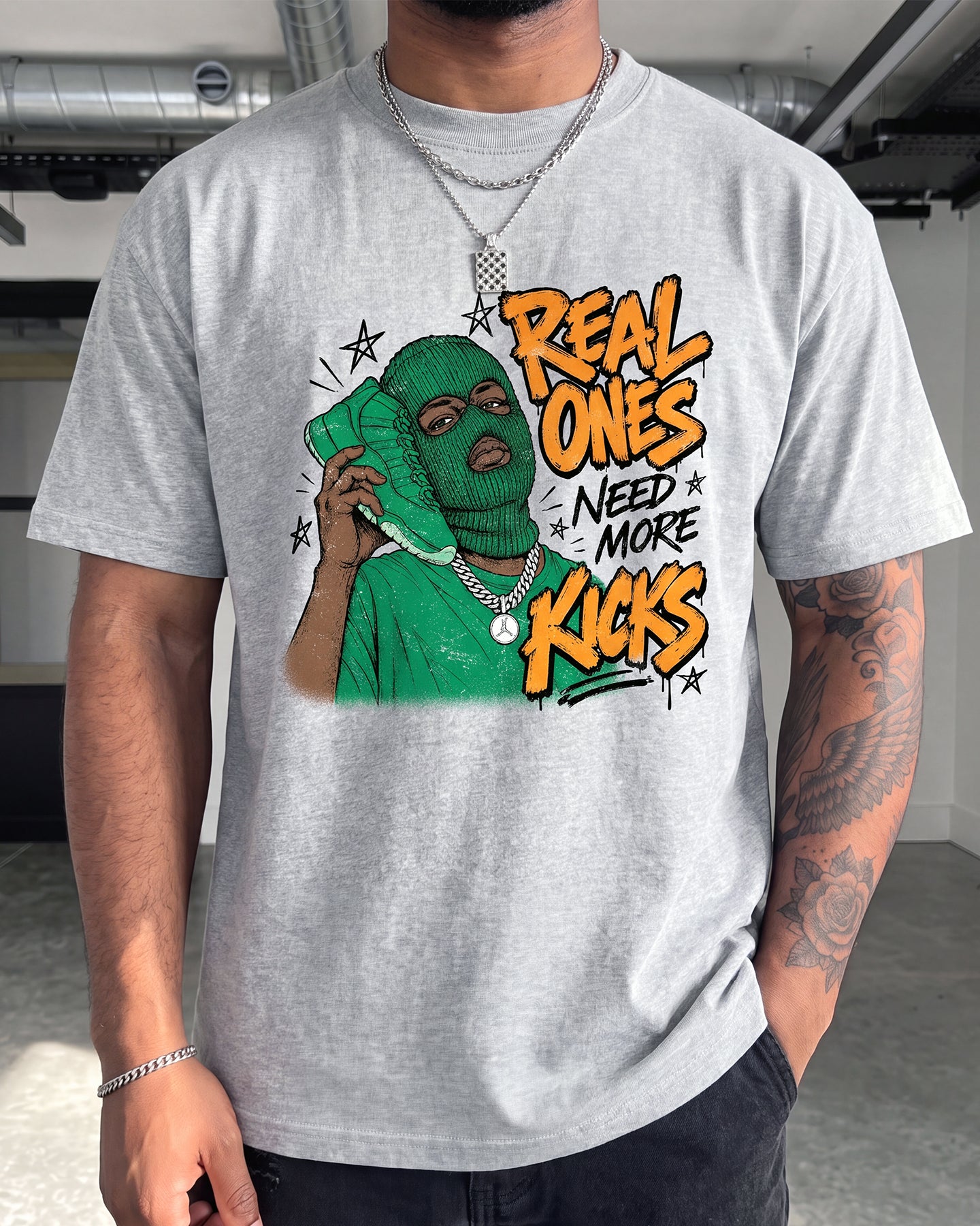 A man wearing a light gray heather t-shirt with a colorful graphic print. The graphic features a green-masked figure holding a phone and bold text that reads 'REAL ONES NEED MORE KICKS' in orange and black.