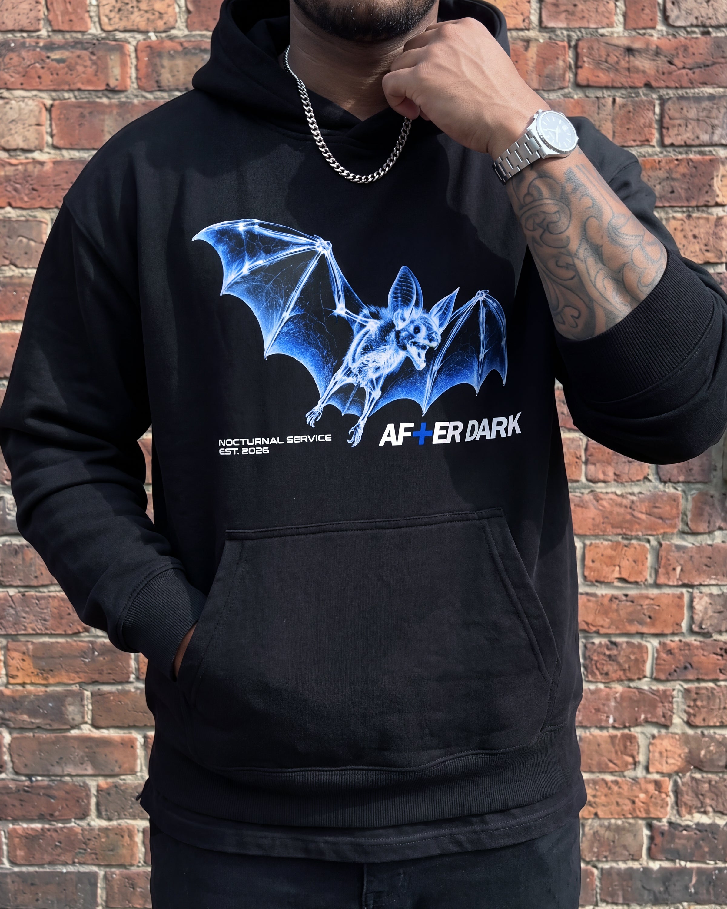 A man wearing a black hoodie with a blue bat graphic and the text 'AFTER DARK' and 'NOCTUNAL SERVICE EST 2025' on the front, standing against a red brick wall.